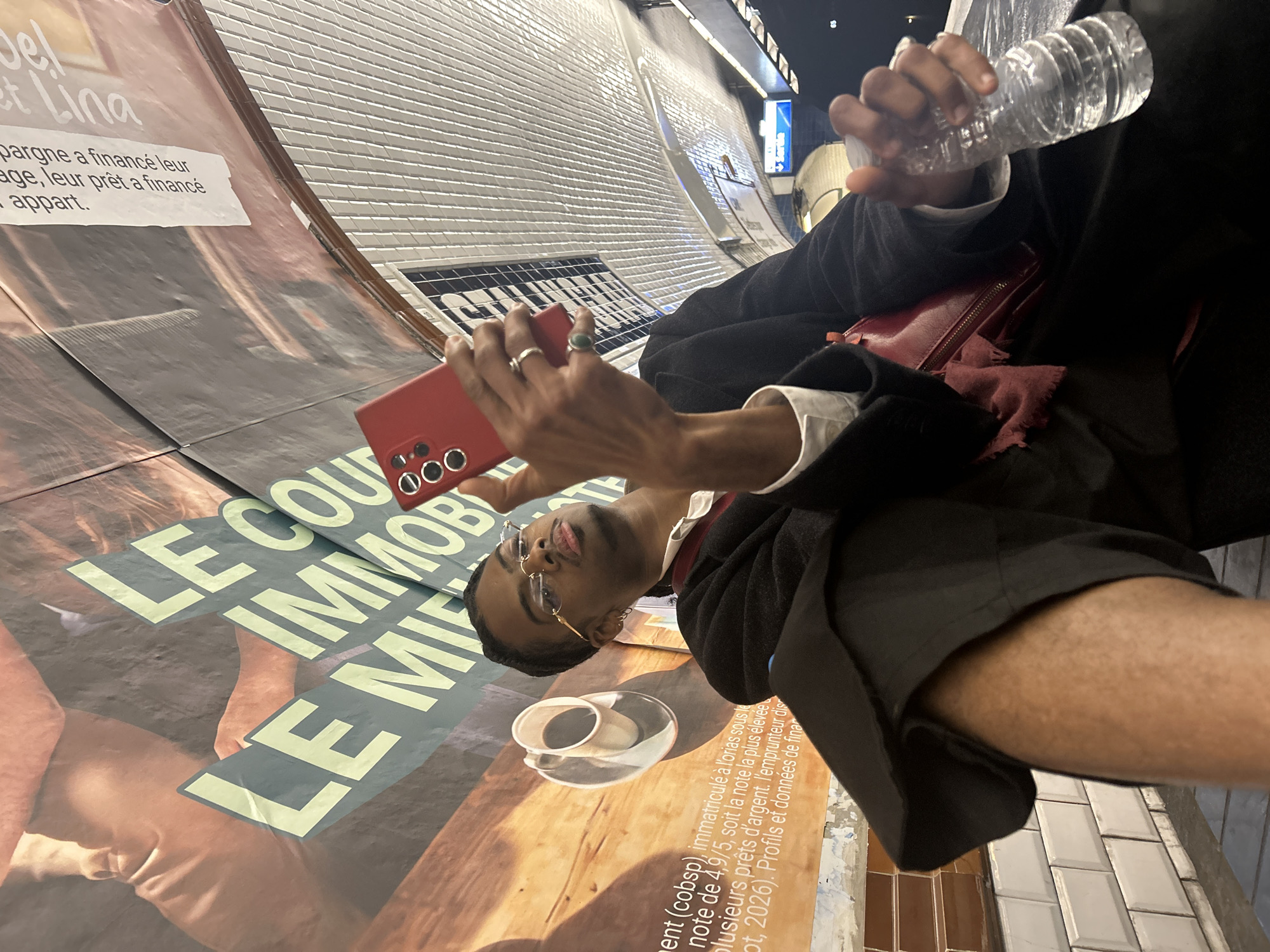 A person stands in a Paris Métro station, holding a red phone in their right hand and a water bottle in their left. They are facing an advertisement poster affixed to the curved wall of the station tunnel. The advertisement includes the phrases "LE COU" and "IMMOB" in large lettering, alongside a cup of coffee. The person is wearing eyeglasses, a dark jacket, and a white-collared shirt, and they have a reddish bag on their person. The station's tiled wall is visible in the lower-right corner.