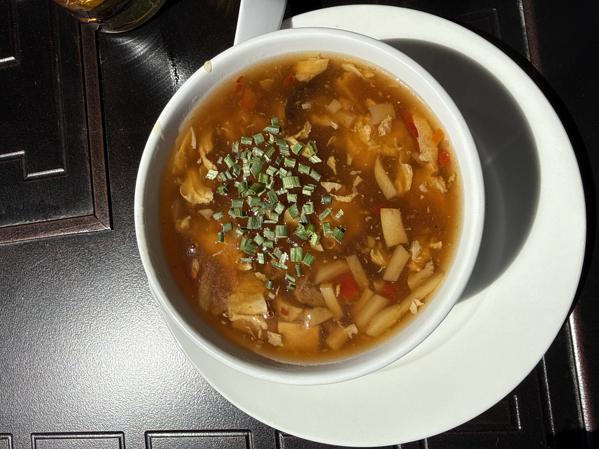 A close-up, high-angle shot captures a steaming bowl of soup, likely hot and sour soup, served on a dark, textured table surface. The soup, a rich amber-brown, fills a white ceramic bowl and is generously garnished with finely chopped green chives or scallions scattered across its surface.
Within the hearty broth, numerous ingredients are visible, including irregular pieces of shredded yellow egg, rectangular slices of light-colored bamboo shoots, and small cubes that appear to be tofu. Slender, bright red strips, possibly bell pepper or chili, add a touch of color to the mixture.
The white bowl rests on a slightly larger white plate or saucer, which casts a prominent, dark shadow across the table surface due to strong, direct lighting. A white ceramic spoon handle is partially visible at the top left, resting in the bowl. The table or placemat beneath is dark grey or black with a distinctive, slightly pebbled texture, and features a subtle raised pattern along its visible edges. In the upper left corner, a portion of a glass containing a golden-brown liquid, possibly beer or tea, is partially visible.
The scene is brightly lit by what appears to be direct sunlight, casting sharp, defined shadows and highlighting the texture of the table surface, suggesting a daytime setting, possibly outdoors or near a window in Hainburg an der Donau, Austria. No people or visible text are present in the image.