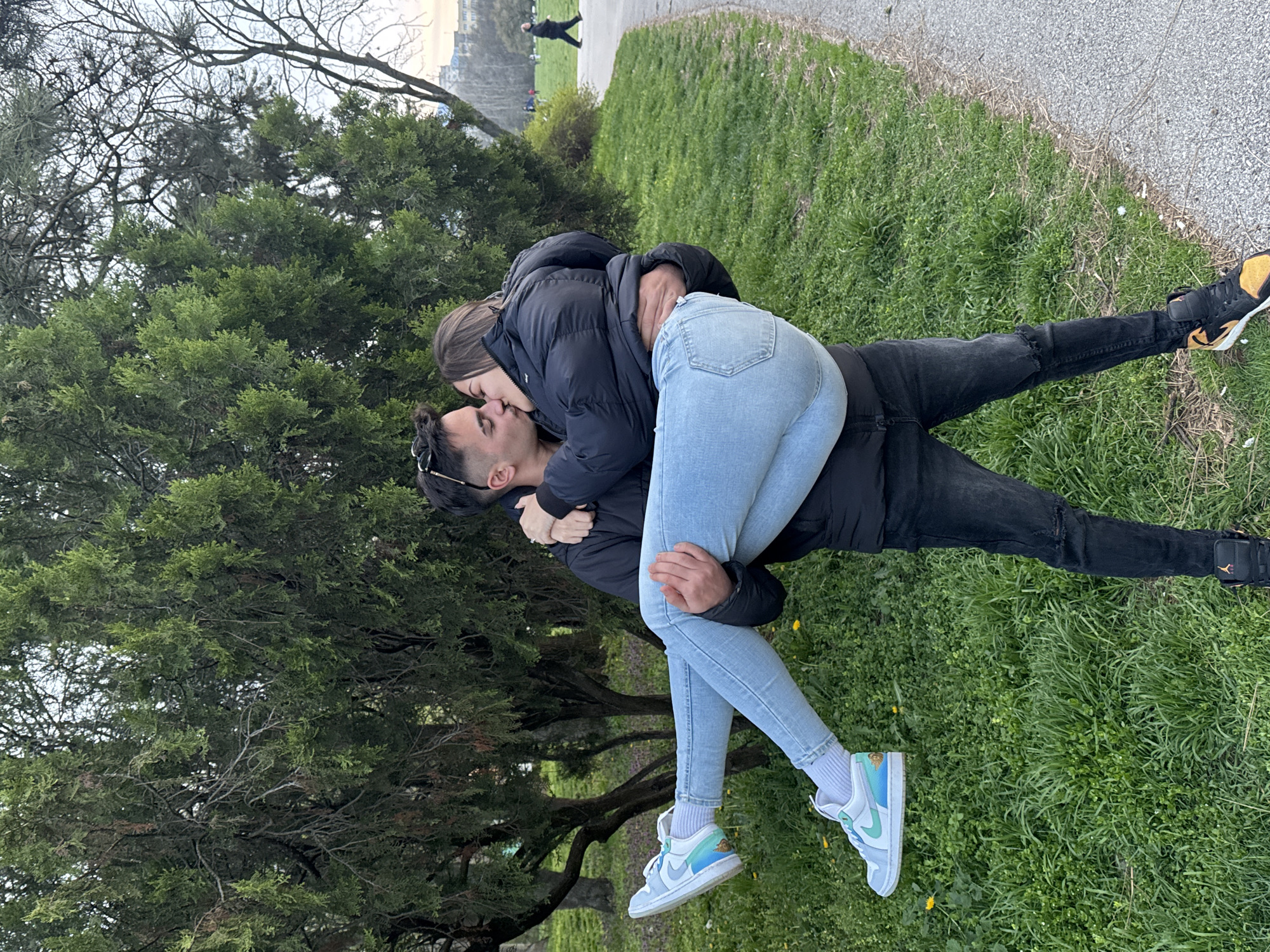 A photograph depicts a man and a woman in an intimate embrace, kissing. The woman is wearing a dark puffer jacket and light blue jeans, with light blue sneakers. The man is wearing a dark jacket and dark jeans with yellow and black sneakers. The setting appears to be a park or green space, with trees and grass visible. In the background, a path is visible with a person walking along it. The image suggests a public display of affection in an outdoor environment. The location is most likely Sofia, Bulgaria.