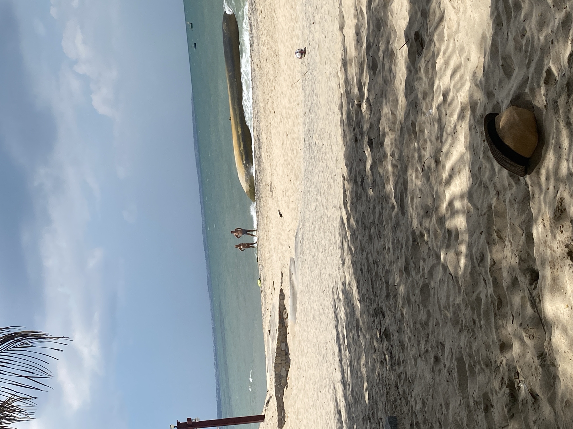 Two individuals, one male and one female, stand on a sandy beach near the water's edge. In the background, a capsized vessel, appearing to be a boat or canoe, rests partially submerged in the ocean. The sky is partly cloudy, with patches of blue visible. The foreground of the image is dominated by sand, with shadows cast from unseen foliage. A straw hat rests on the sand in the lower right corner, and a partially deflated ball is also visible on the sand. The scene appears to be a beach in Phu Thuy Ward, Vietnam, characterized by its sandy terrain and the presence of the ocean.