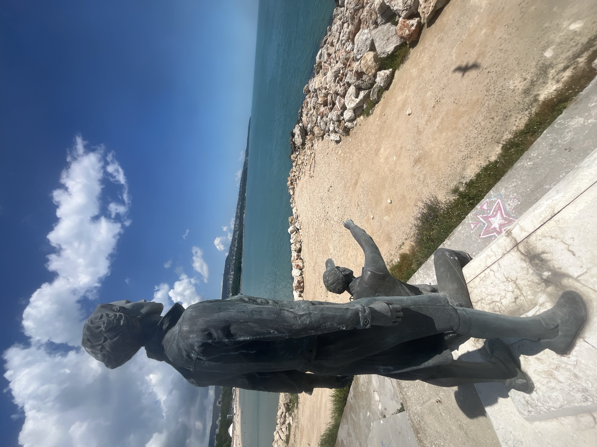 Two bronze statues are positioned on a stone plinth overlooking the sea. The foreground statue depicts a standing figure with short, curly hair, wearing a jacket and trousers, looking out towards the horizon. To the right and slightly lower, a second statue shows a figure kneeling, pointing towards the sea. The background features a sandy embankment with a rocky seawall, the turquoise waters of the Black Sea, and a distant coastline with buildings and a beach. The sky is blue with scattered white clouds. The scene appears to be located in Varna, Bulgaria, likely at the Sea Garden promenade, a popular tourist area with public art installations.