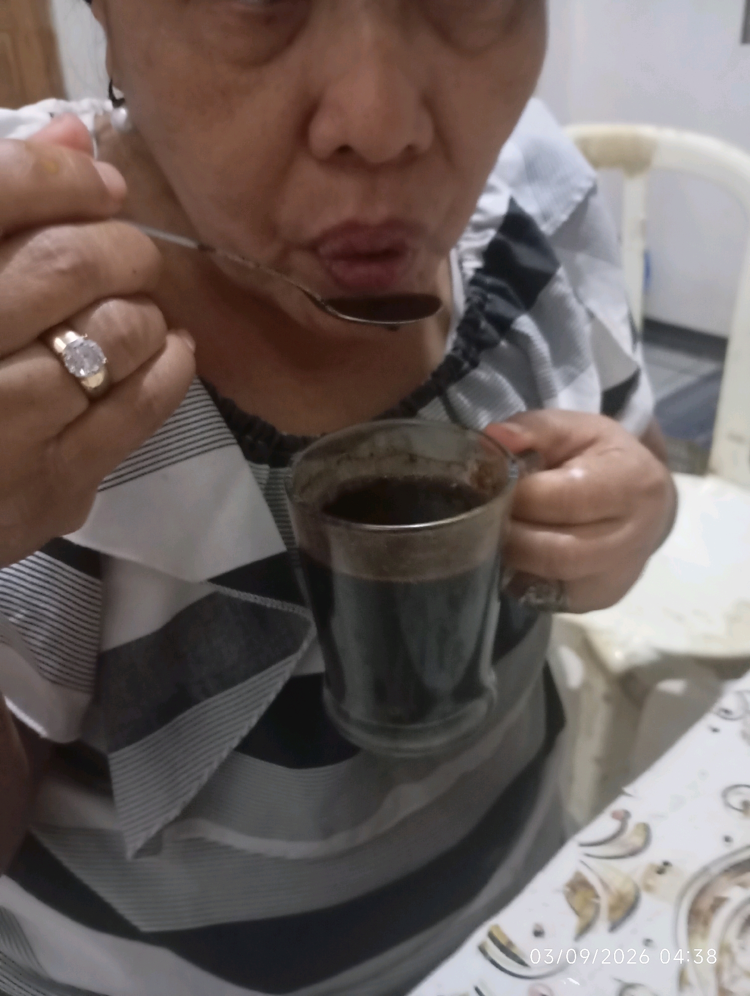 A medium shot shows a person drinking from a spoon, holding a mug of coffee. The person is wearing a striped shirt. A ring is visible on their left hand. The background is out of focus, showing part of a chair and a table with a patterned surface. A timestamp reads "03/09/2026 04:38". The image is undated, and there is no indication of the specific location within Klecorejo, Indonesia.