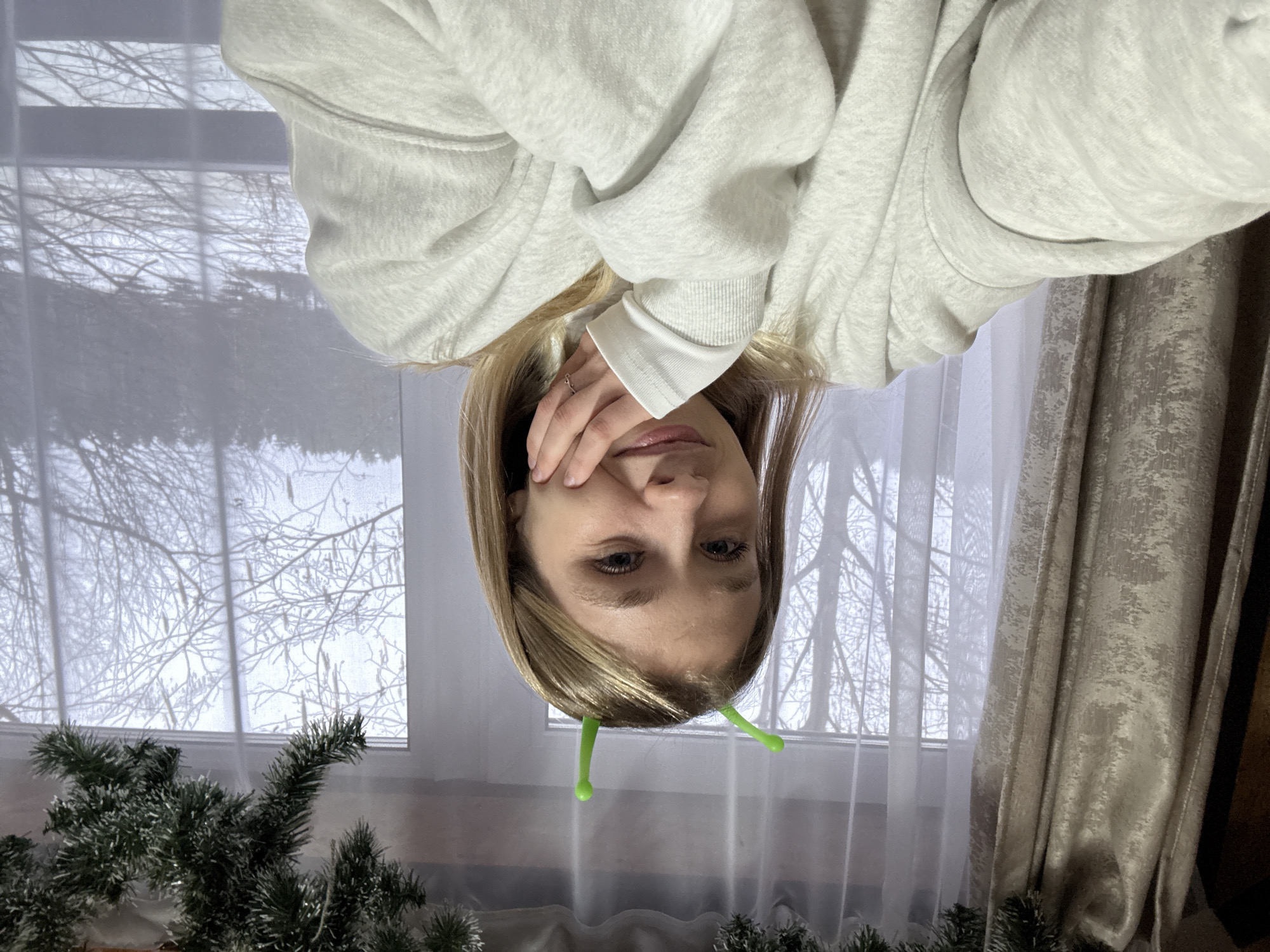 A woman with blonde hair and a light grey hoodie is in the frame, appearing to be upside down. She is holding her hand to her face. She is wearing green antennae. There is a window in the background with bare tree branches visible outside. There is also a curtain and a green garland in the frame. There is no evidence in the image to suggest it was taken in the city of Прикамский, Russia.