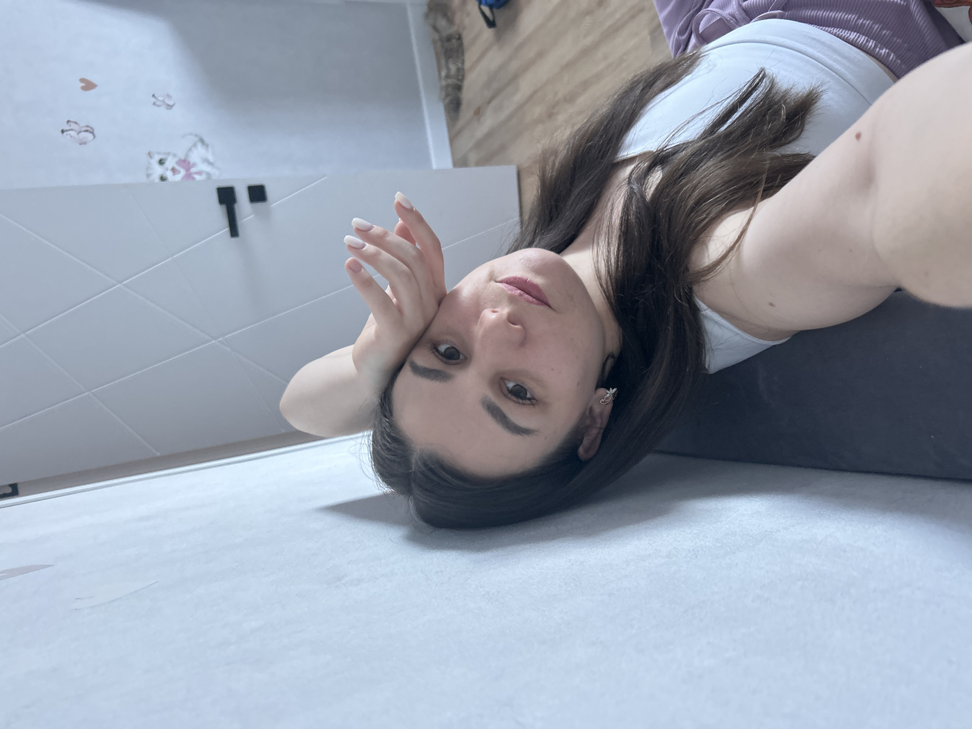 A selfie shows a woman with long brown hair wearing a white tank top and a purple shirt, lying on a light gray surface. Her head is tilted, with her right hand near her face. In the background is a white wall with a textured pattern and cartoon-style stickers. Visible above the wall are sections of a wooden floor. Based on metadata (not present in the images but provided to the analysis), the location is Chaikovsky, Russia.