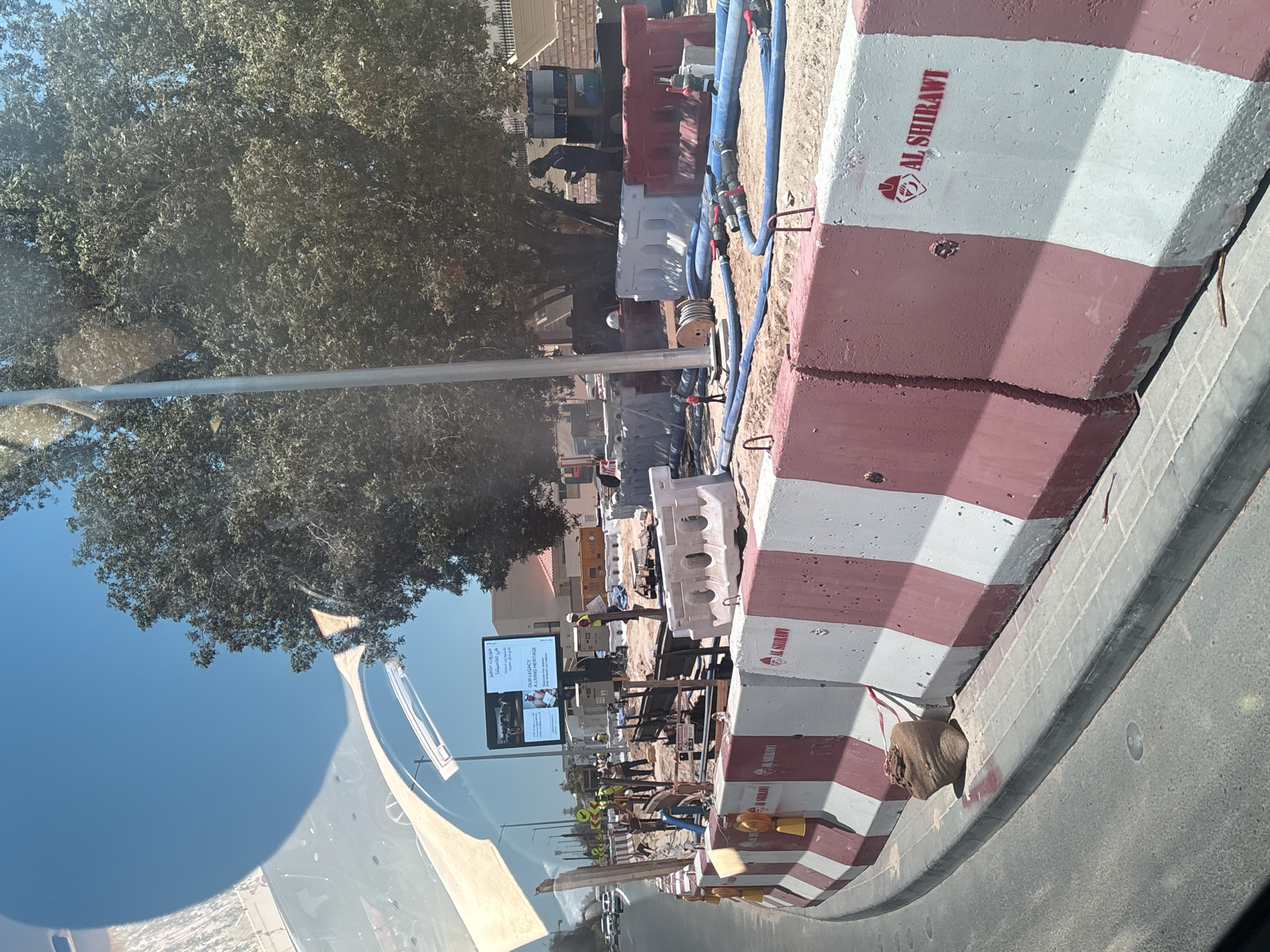The image shows a construction site in Dubai, United Arab Emirates, with concrete barriers marked "AL SHIRAWI" lining the road. Construction workers in safety vests are visible, along with heavy machinery and large blue hoses. A digital billboard displaying the text "OUR LEGACY, A LIVING HERITAGE" is also present. The scene depicts ongoing infrastructure work under a clear blue sky, with lush green trees in the background.