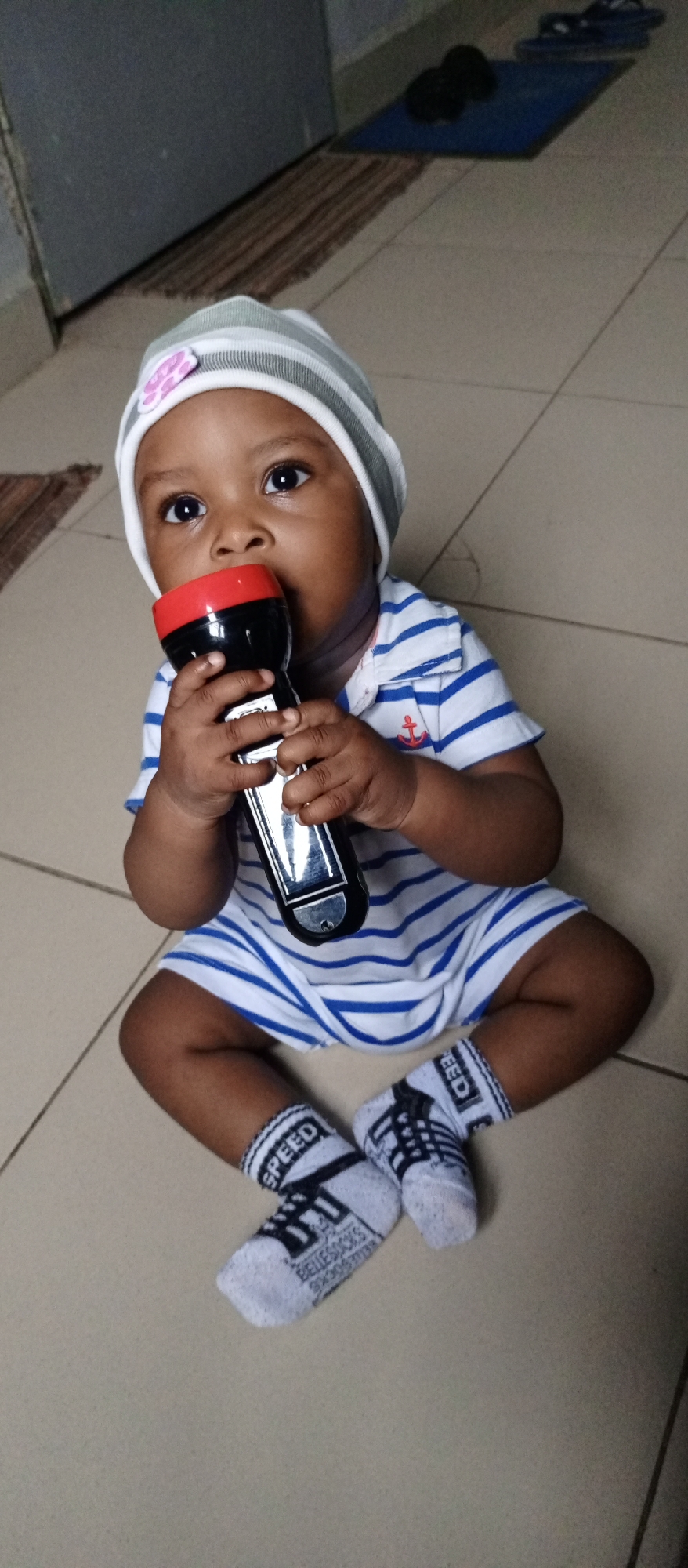 A baby sits on a tile floor in an indoor space. The baby wears a striped hat with a flower applique, a blue and white striped sailor suit, and socks with the word "SPEED" printed on them. The baby is holding a flashlight to their mouth. In the background, there's a door and a small blue mat with dark objects on it. The location is Unknown, Unknown.