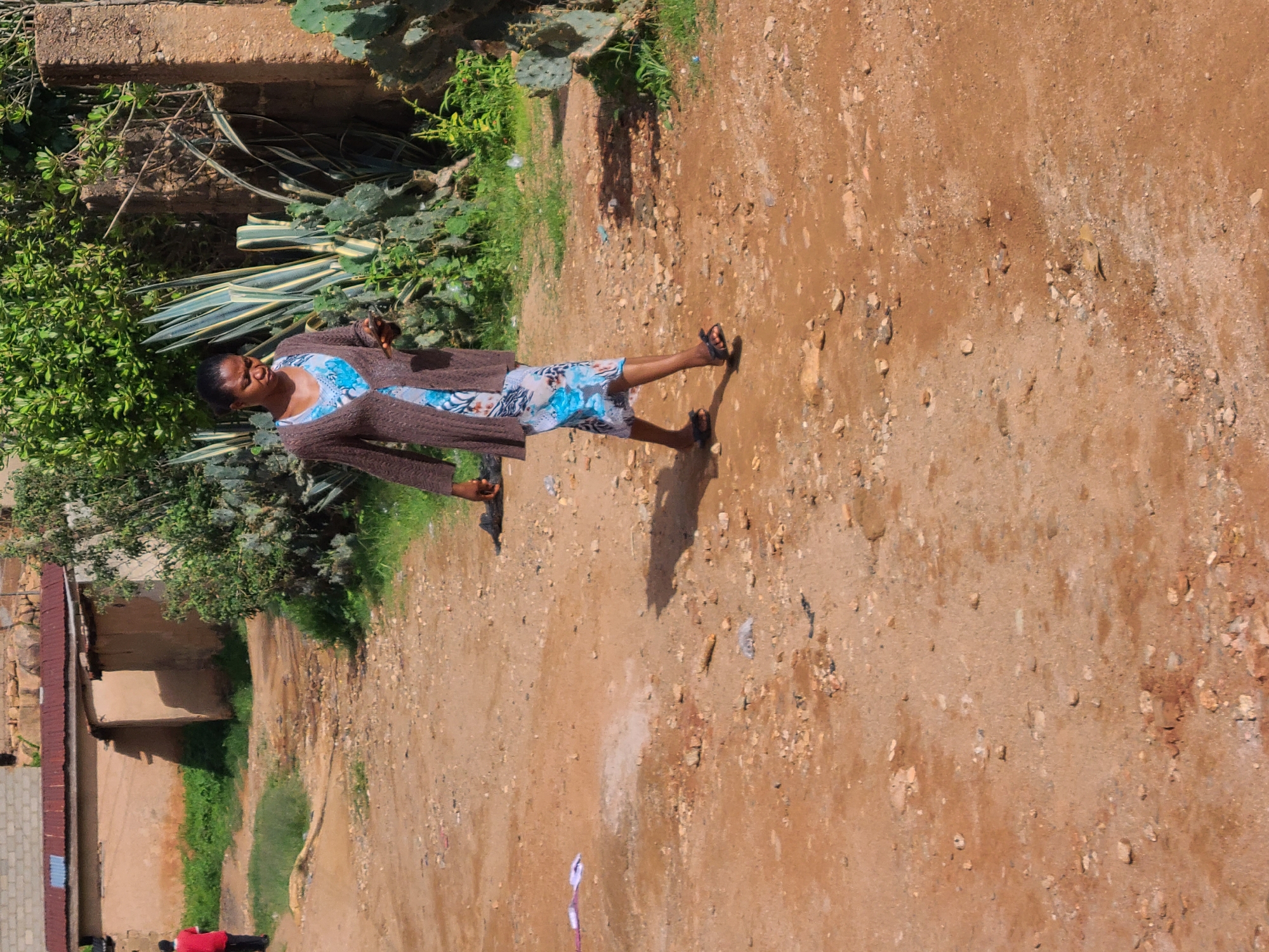 A dark-skinned woman walks on a dirt road in a rural area. She is wearing a long, brown cardigan over a blue and white patterned dress. She is holding a mobile phone in her right hand. The road is covered in dirt and small rocks. In the background, there are several buildings with red roofs, some green bushes, and cactus plants. The scene appears to be a sunny day. The location is described as Jos, Nigeria.