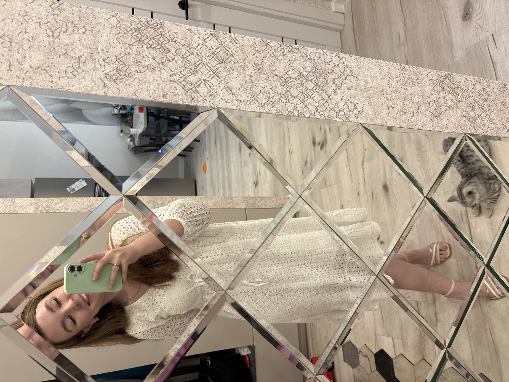 A woman is taking a selfie in a mirror. She is wearing a white eyelet dress and gold and white sandals. She is holding a light green smartphone in her right hand. A tabby cat is lying on the floor in the lower right corner of the image. The mirror is made of square and diamond-shaped panels. The background of the image shows a room with light wood floors and a table with chairs. The scene appears to be an indoor residential setting, possibly an apartment or house in Izhevsk, Russia.