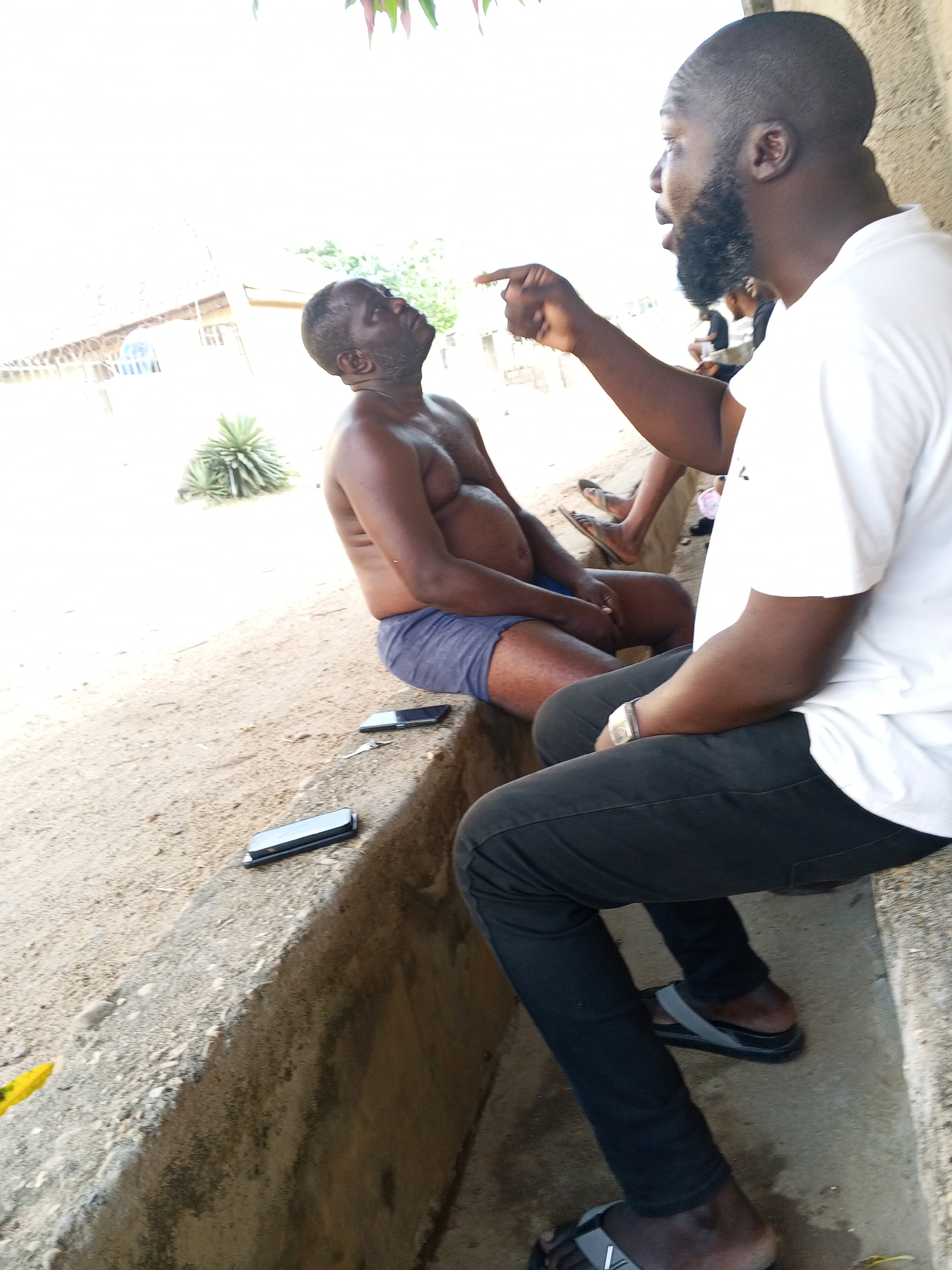 Two men are central to an outdoor scene, appearing to be engaged in a lively conversation. On the right, a man with a beard and a white t-shirt sits with black pants and sandals, actively gesturing with his right index finger while speaking. A watch is visible on his left wrist. Facing him, on the left, an older man sits shirtless, wearing blue shorts, looking upwards and to the right, seemingly listening intently or reacting to the speaker. He has a prominent belly.
Both men are seated on a low concrete ledge or step. Two black smartphones are visible on the ledge between them. In the background, other individuals are partially visible, suggesting a small gathering. The setting appears to be outdoors, with dusty ground and some distant, indistinct structures, possibly with simple roofing. A succulent plant is visible behind the shirtless man. The lighting is very bright, indicative of daytime, with significant overexposure in the upper left and background sky, suggesting a sunny day. No discernible text is visible anywhere in the image. The environment is consistent with an outdoor, possibly casual, setting in a location like Adonkolo, Nigeria.