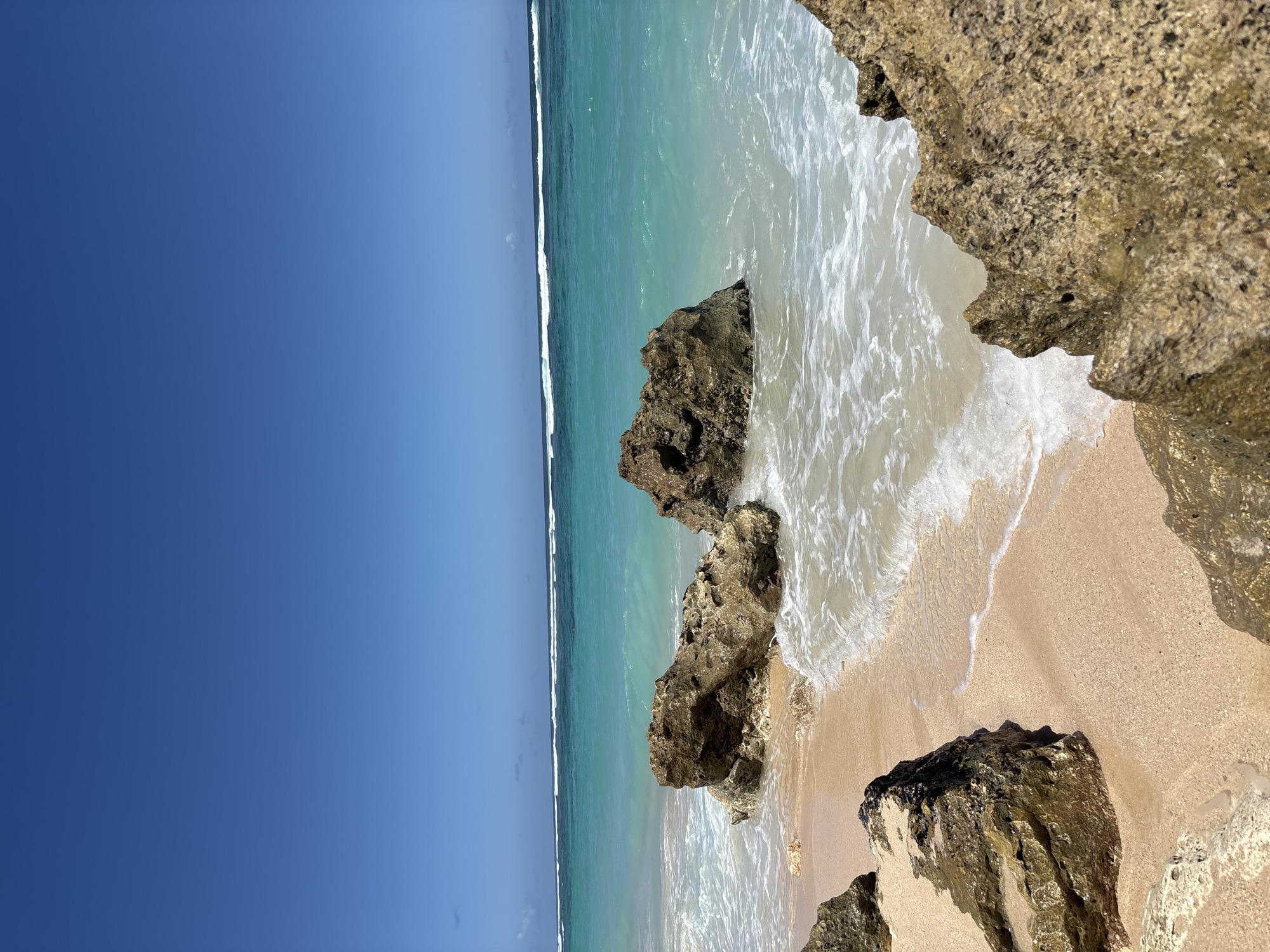 The image depicts a tropical beach scene in Kuta Selatan, Indonesia. The focus is on a sandy beach where waves gently lap against the shore. Several rocky outcrops of a light brown color are partially submerged in the water. The sea transitions from a light turquoise near the shore to a deeper blue further out, meeting a clear, deep blue sky. The lighting suggests it is a bright, sunny day. No people are visible in the scene. The overall impression is of a natural coastal landscape.