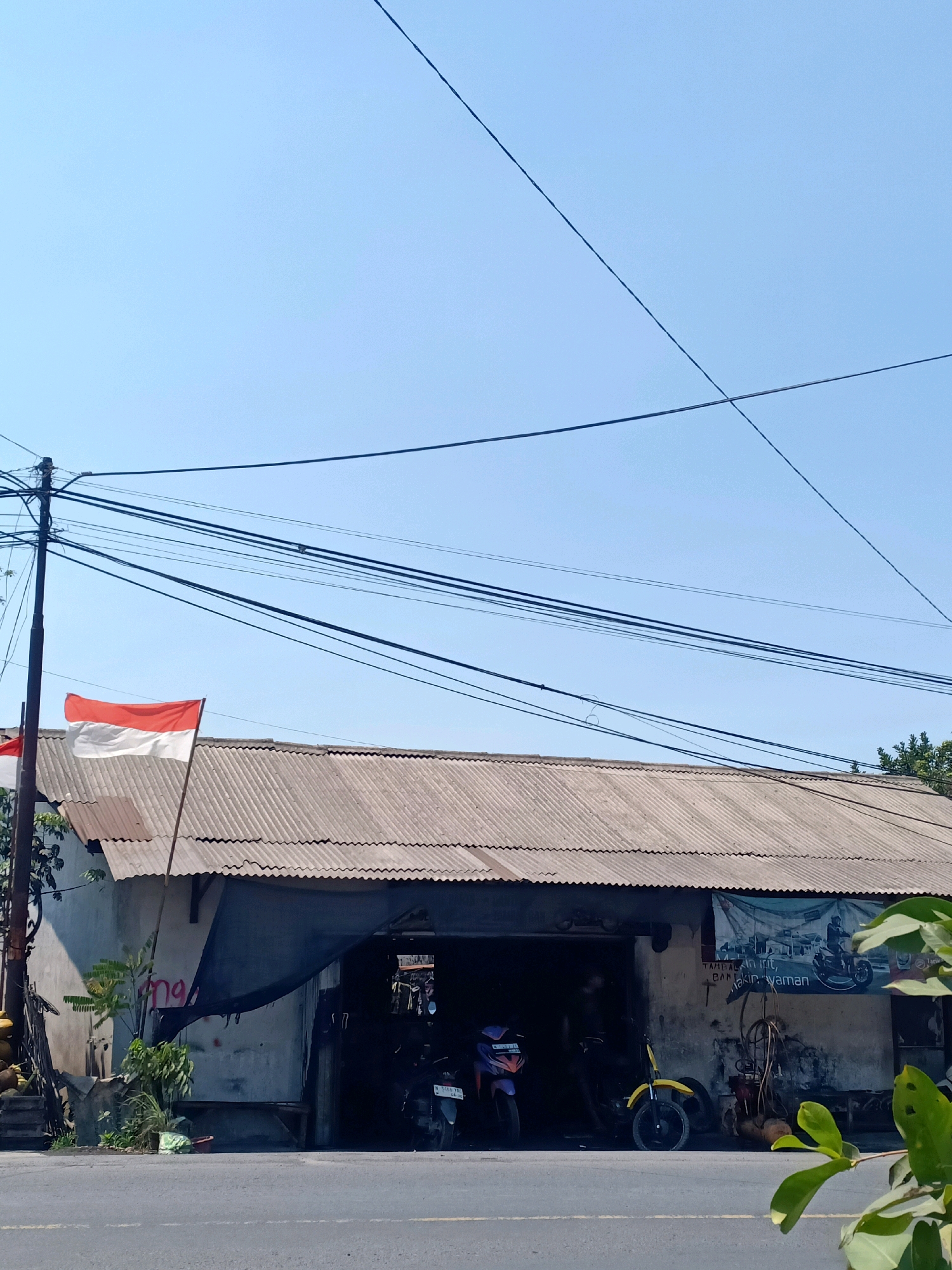 The image depicts a scene in Jarit, Indonesia, featuring a small, open-fronted building that appears to be a workshop or roadside stall. The building has a corrugated metal roof and concrete walls. An Indonesian flag, with red and white stripes, is visible flying from a pole to the left of the entrance.
In front of the building, a paved road runs horizontally across the lower portion of the frame. Several motorcycles are parked inside and near the entrance of the building, suggesting it is a place that services or sells them. One motorcycle in the foreground is purple and black, while a yellow motorcycle is visible to the right of the entrance.
A banner with an image of a person riding a motorcycle is hung on the right side of the building. Overhead, a complex network of electrical wires stretches across the sky, supported by utility poles. The sky is clear and blue, indicating a sunny day. Lush green foliage is present on the left side of the building and in the lower right corner of the frame. The overall impression is of a typical, utilitarian roadside establishment in a tropical or subtropical urban or semi-urban environment.