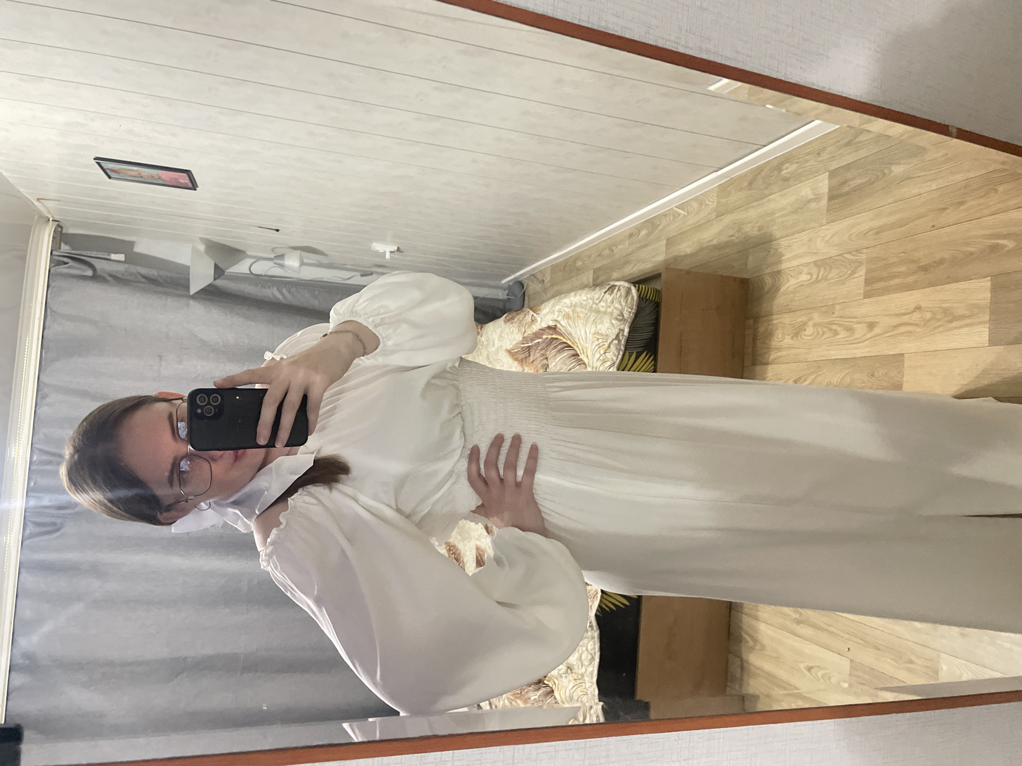 A young woman with brown hair, wearing glasses and a white dress with puff sleeves, is taking a mirror selfie in a room. The dress has a smocked waist and a long, flowing skirt with a slit. She is holding a smartphone in her right hand. In the background, a bed with a patterned comforter is visible, along with a floor lamp and a framed picture on the wall. The room appears to be a bedroom, likely in a residential building in Сырково, Russia. The floor is made of light-colored wood planks.