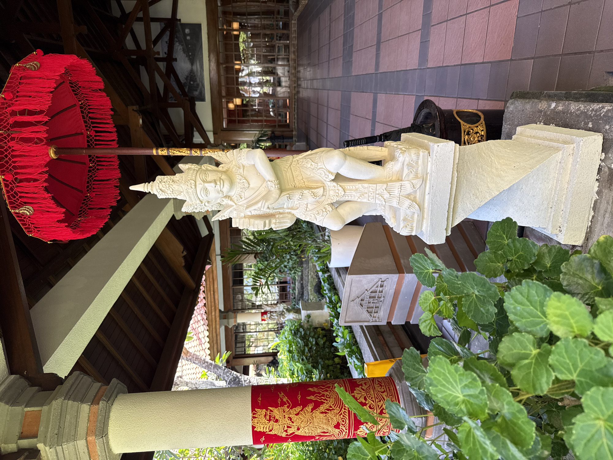 A horizontal shot of a statue and courtyard. The statue is of a figure in traditional Balinese garb, laying down. It is white, on a tall white pedestal. A red umbrella is to the left of the statue, decorated with a gold design on the bottom. To the right is lush green vegetation. In the background, there is a courtyard with trees and buildings. The setting suggests a tourist-friendly location, characteristic of Kuta Selatan, Indonesia.