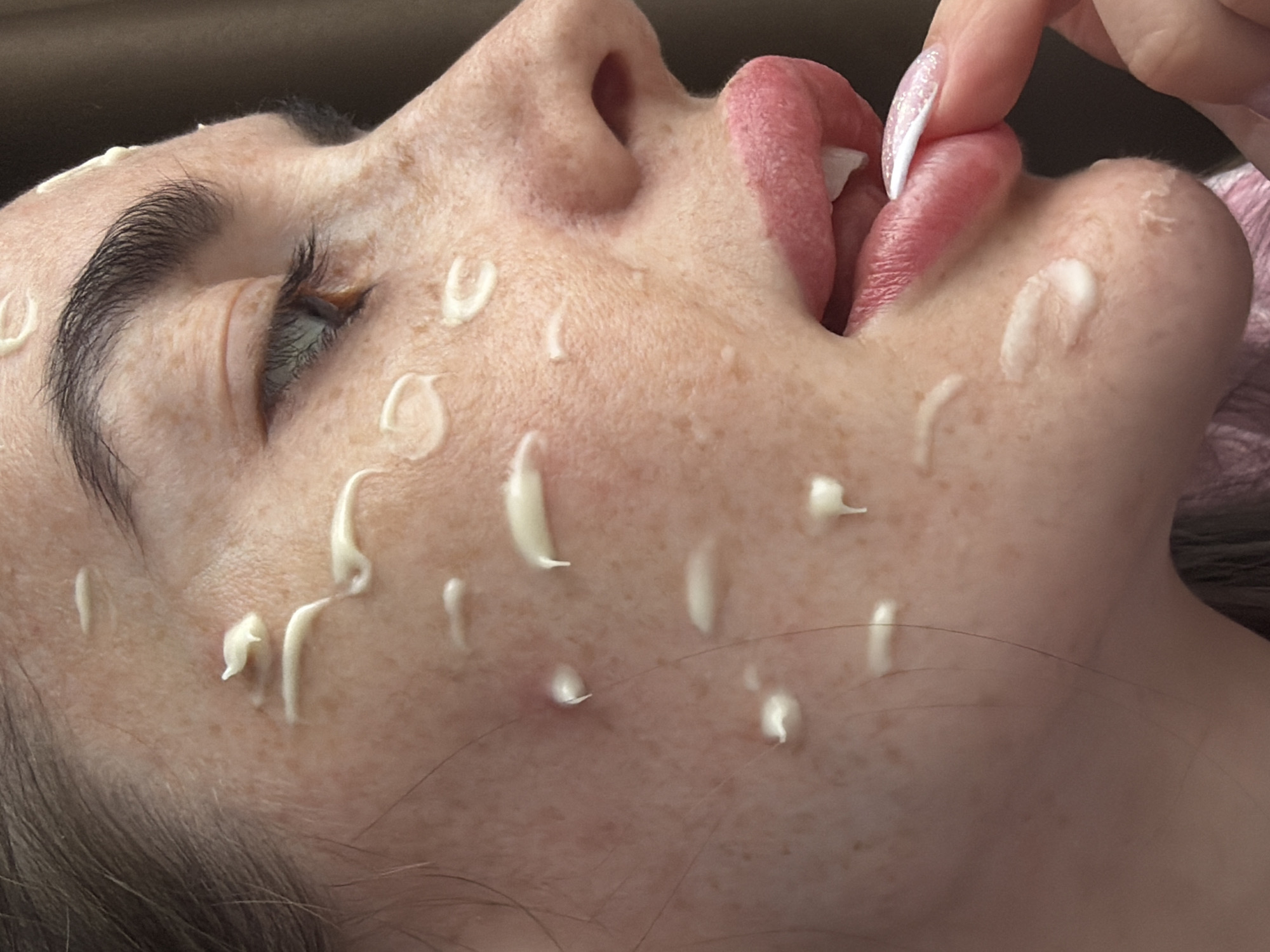 A close-up, angled view focuses on a fair-skinned person with freckles. Their face is smeared with small dollops of a white substance. The person's lips are slightly parted, with the tongue partially protruding. One of their fingers, adorned with a white and glittery nail, touches their lip. The background is blurred and indistinct. The location and country are not discernible from this image.