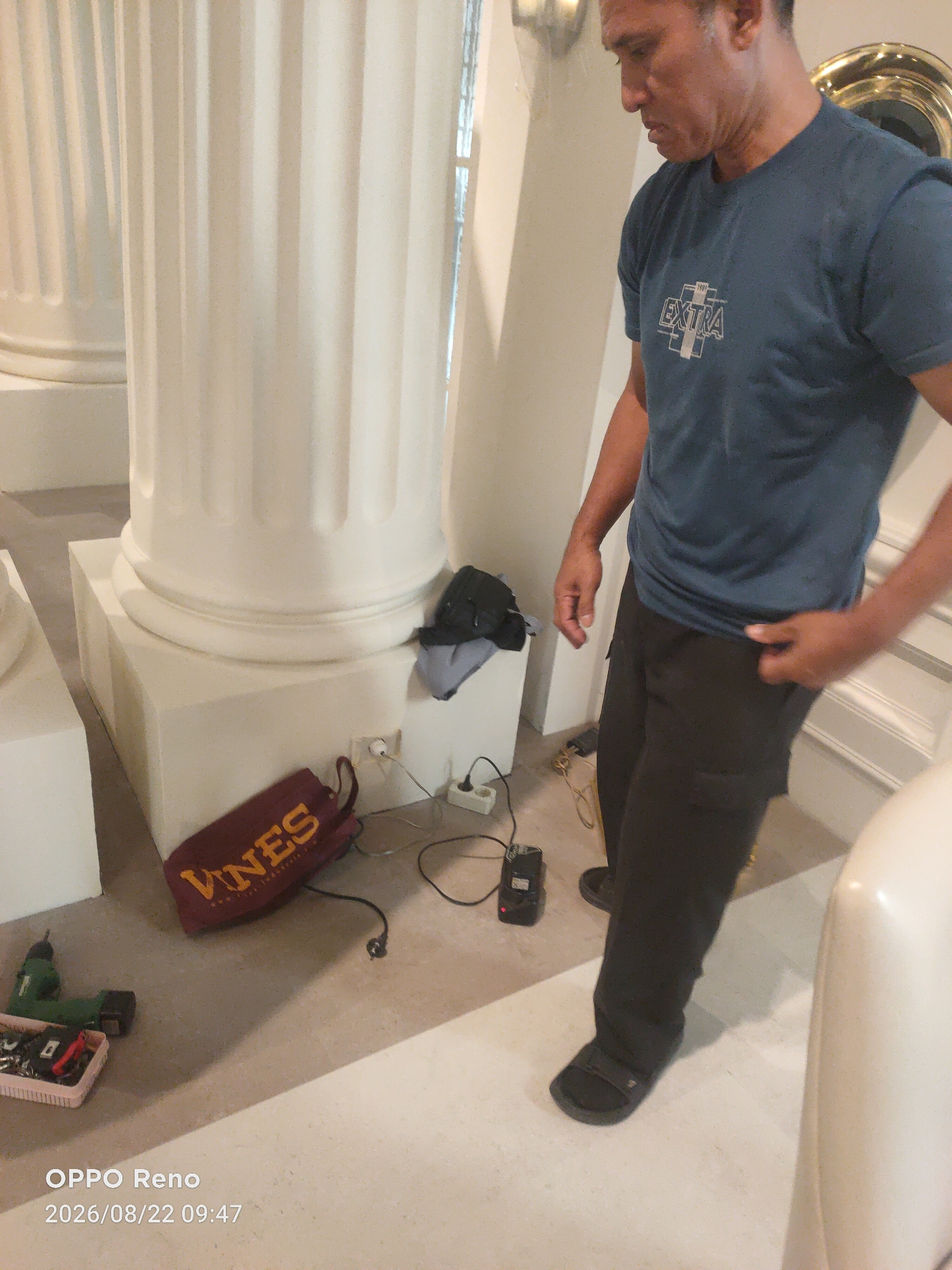An adult male wearing a dark blue t-shirt with "EXTRA" printed on the front and dark pants stands next to a fluted white pillar in what appears to be an indoor setting. A maroon bag with "VINES" written in yellow lettering is on the floor near the base of the pillar. A green power drill and a pink plastic container filled with various tools are to the left of the bag. Several electrical cords and a small black device are also visible on the floor. The background shows more of the white pillar and a wall. The lighting suggests an indoor environment. The timestamp "OPPO Reno 2026/08/22 09:47" is present in the lower left corner of the image.