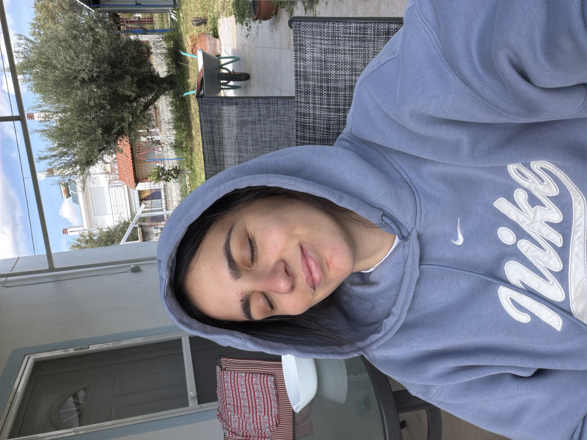 A woman in a light blue Nike hooded sweatshirt is sitting outside in Παραλία Οφρυνίου, Greece. She has her eyes closed and appears relaxed. She is sitting in a chair with a grey mesh back. A glass table is in front of her, partially covered with a red and white striped cloth and a white object. In the background, there is a tree, a house with a white facade and orange roof tiles, and a wheelbarrow. The sky is visible with some scattered clouds.