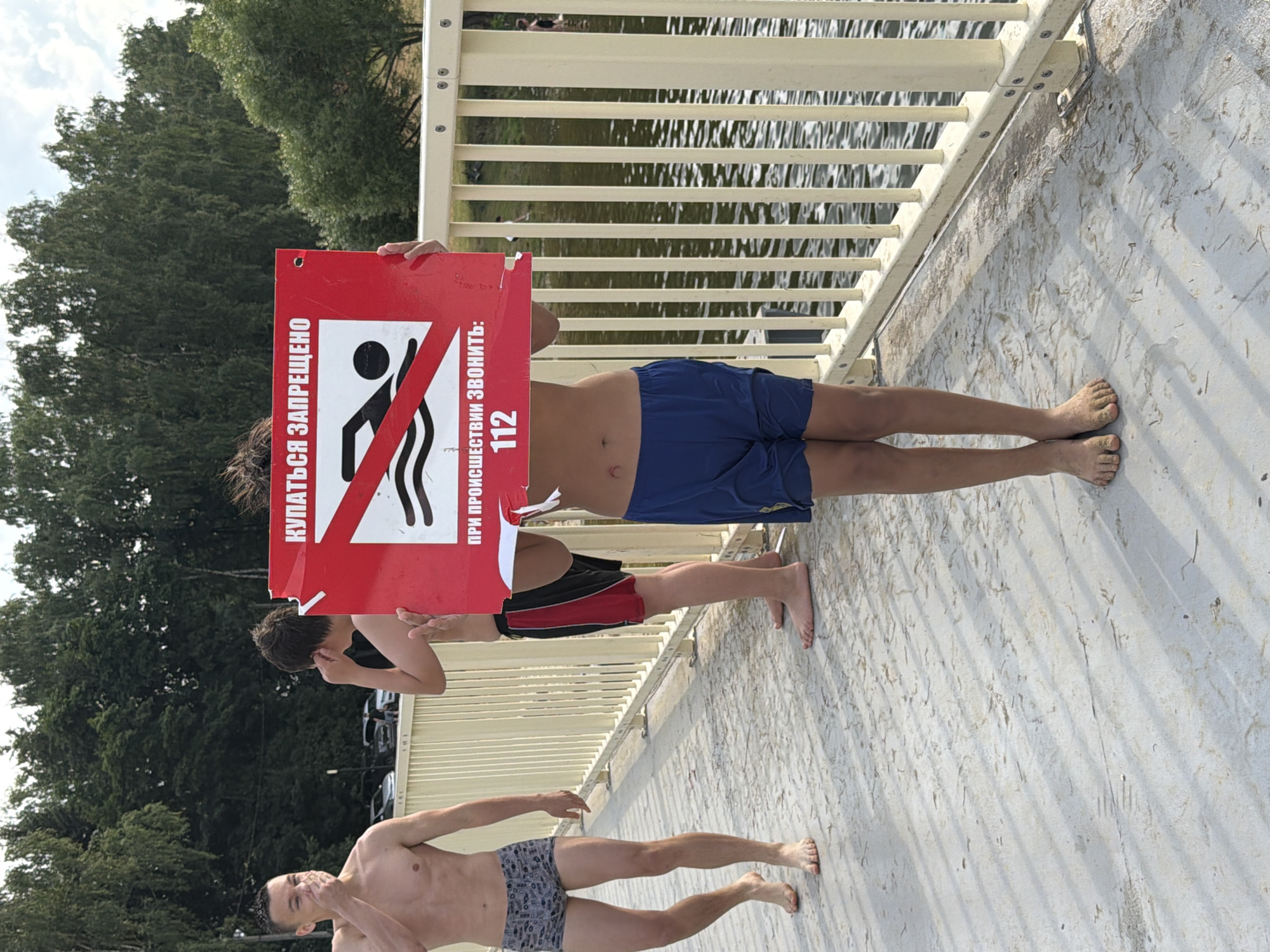The image shows three young men on a concrete walkway next to a body of water, likely a river or lake, under a bright sky. The perspective is from above, looking down.
The two men in the foreground are shirtless and wearing swim trunks. The man at the bottom is facing away from the camera, showing his patterned swim trunks. The man above him is also facing away, wearing black and red swim trunks. He is looking back and appears to be gesturing or speaking.
A third young man, in the middle ground, is holding up a red sign with a white prohibition symbol on it. The symbol depicts a person swimming, crossed out. The sign has Russian text. The man holding the sign is shirtless and wearing blue swim trunks. He is looking down at the sign.
The walkway has a light-colored surface, possibly concrete or stone, with some debris scattered on it. A metal railing runs along the edge of the walkway, separating it from the water. The water is rippling and reflects the sunlight. Trees with green foliage are visible in the background, suggesting a natural or park-like setting. The sun appears to be high in the sky, indicating it's daytime.
The visible text on the sign reads "КУПАТЬСЯ ЗАПРЕЩЕНО" which translates to "SWIMMING IS PROHIBITED". Below that, it says "ПРИ ПРОИСШЕСТВИИ ЗВОНИТЕ: 112", meaning "IN CASE OF EMERGENCY CALL: 112". The presence of this sign and the men in swim trunks suggest that swimming is not permitted in this area, despite the proximity to the water. There is no clear indication of distress or urgency among the men.