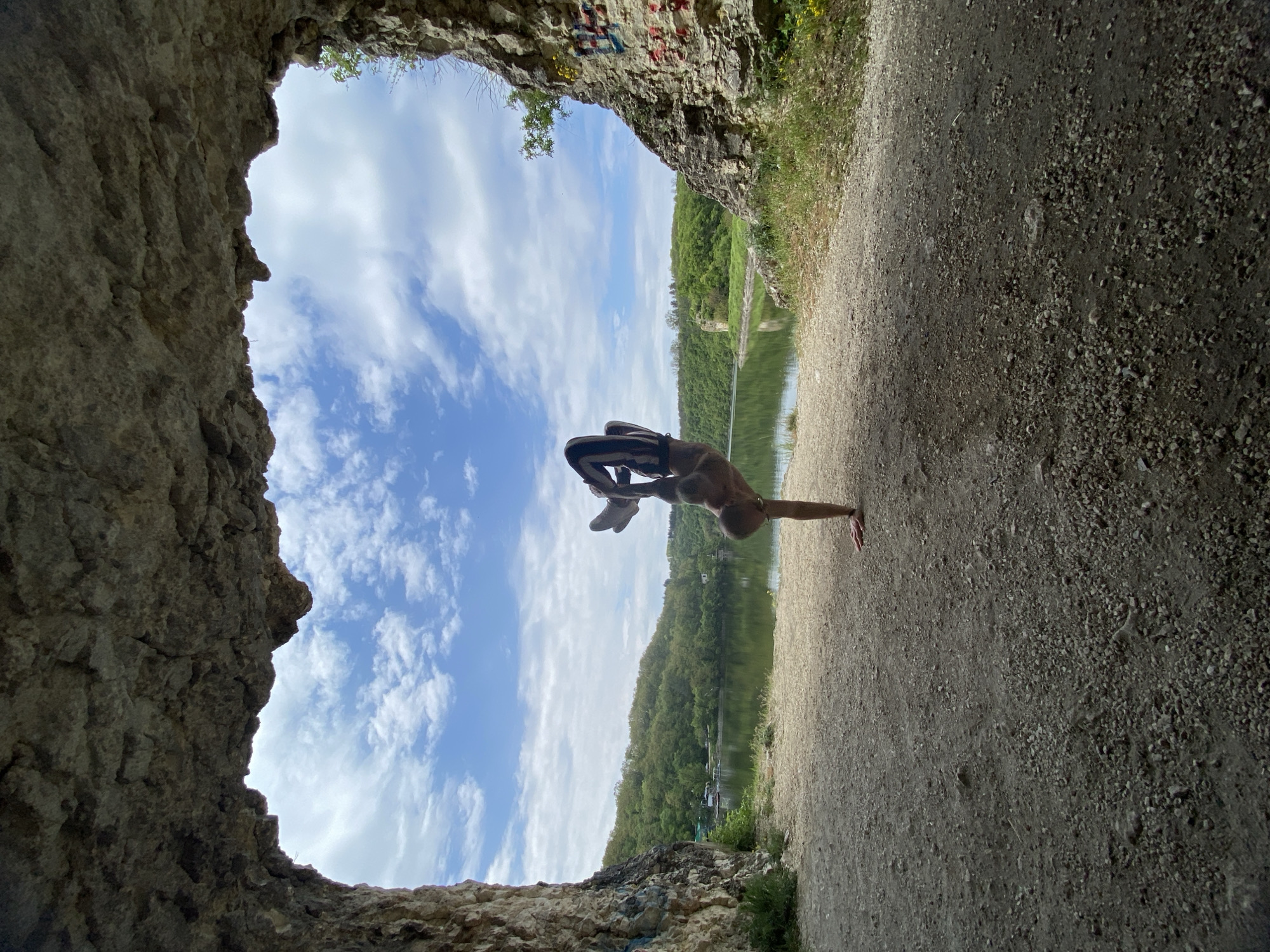 The image presents a view from within a cave-like opening, looking out onto a landscape. Outside the opening, there's a body of water with a tree-lined shore and a blue sky with white clouds. In the foreground, a person is performing a handstand, with their feet pointing upward toward the sky. The cave opening is rough and textured. The ground outside appears to be a dirt or gravel path. Given the request for location specifics, this scene is potentially set within the Kaylaka Park near Pleven, Bulgaria, known for its caves and natural rock formations alongside a lake. Graffiti is visible above the cave entrance.