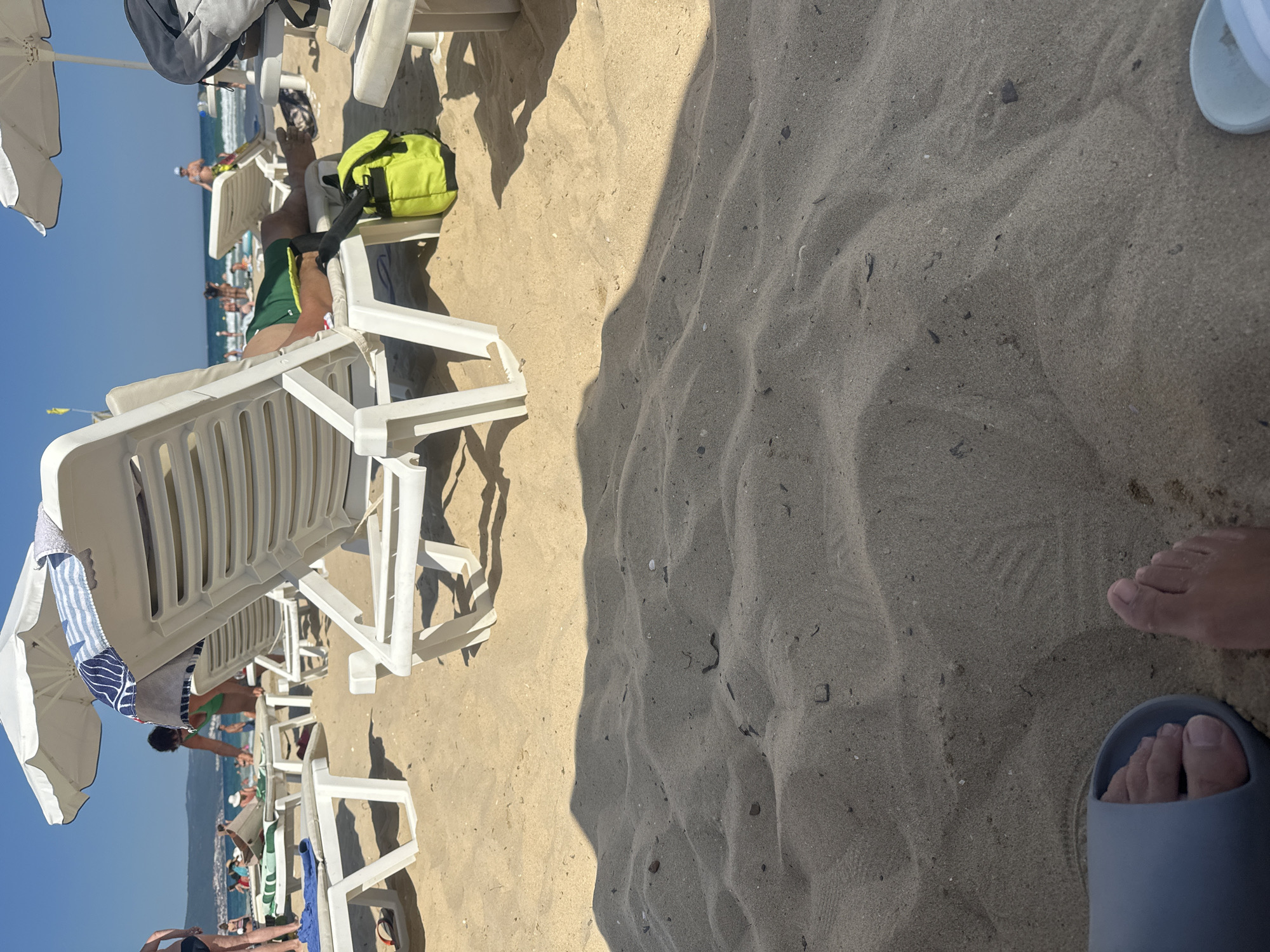 This is an outdoor, eye-level, medium shot of a sandy beach in Nesebar, Bulgaria on a sunny day. In the foreground, a person's bare feet and a light blue sandal are visible on the sand. In the middle ground, several white plastic beach chairs are arranged on the sand. One chair is occupied by a person wearing green shorts, lying down. Another person is partially visible behind a chair, leaning forward. A yellow backpack rests on the sand next to a beach chair. In the background, the sea and a distant coastline are visible under a clear blue sky. Several other people are scattered on the beach and in the water. Umbrellas are set up on the beach. The overall scene depicts a typical summer day at a beach resort.