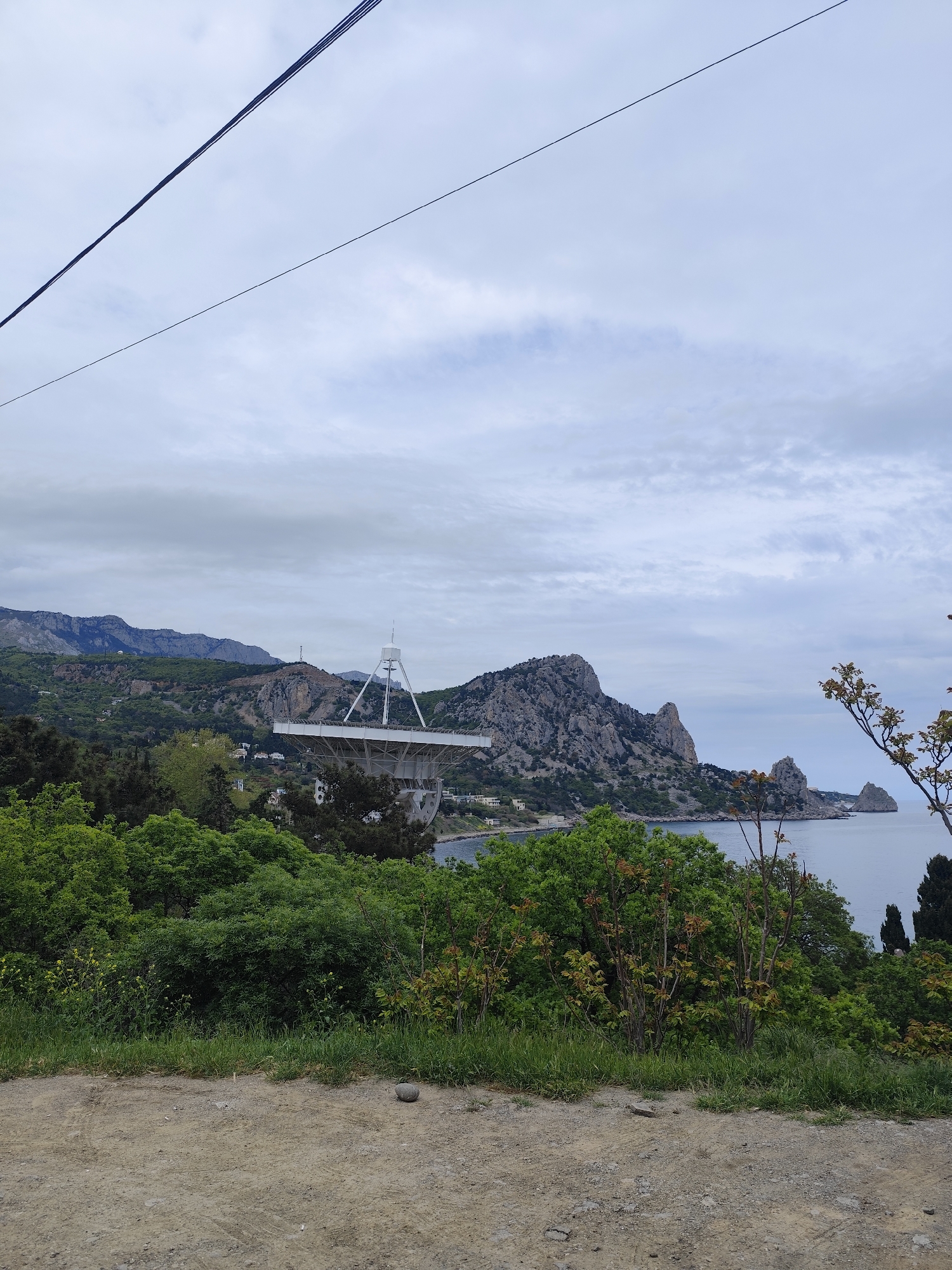 The scene depicts a view overlooking a body of water, likely a bay or coastline, with rocky mountains and a large radio telescope in the middle ground. Green trees and shrubs fill the foreground. The sky is overcast. A dirt road is visible at the bottom of the frame. Power lines stretch across the top of the image. The location is Unknown, Unknown.