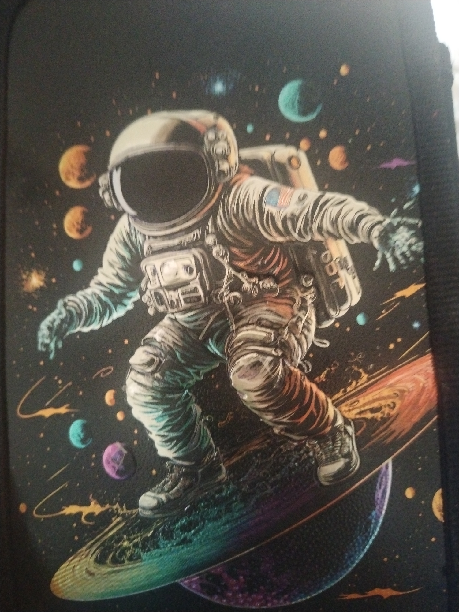 The image depicts an artistic rendering of an astronaut in space, seemingly snowboarding or surfing on a cosmic object. The astronaut is wearing a detailed spacesuit with an American flag patch on the left arm. Various planets, stars, and nebulae are visible in the background, rendered in vibrant colors against a dark, starry expanse. The style of the artwork is illustrative and somewhat stylized, not a photograph. There is no visible context or indication of any location within the city of Tolyatti, Russia, in this image. The image appears to be a print on a dark material, possibly a bag or cover, with a textured surface visible along the right edge.
