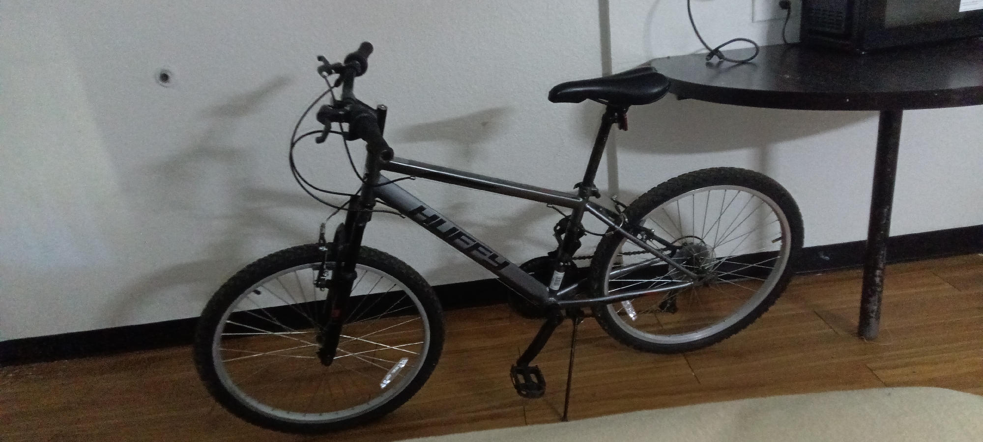 A gray Huffy mountain bicycle is indoors, leaning against a white wall next to a small round black table. The bicycle has black tires, a black seat, and a kickstand. The pedals are black, and the frame has the word "HUFFY" printed on it. The table has a dark surface and a single leg. The floor is wooden, and a light-colored rug is partially visible in the foreground. There are electrical cords and a black object (likely an electronic device) on the table. The scene is likely in Fort Smith, United States of America, but there are no specific landmarks or identifiers to confirm.