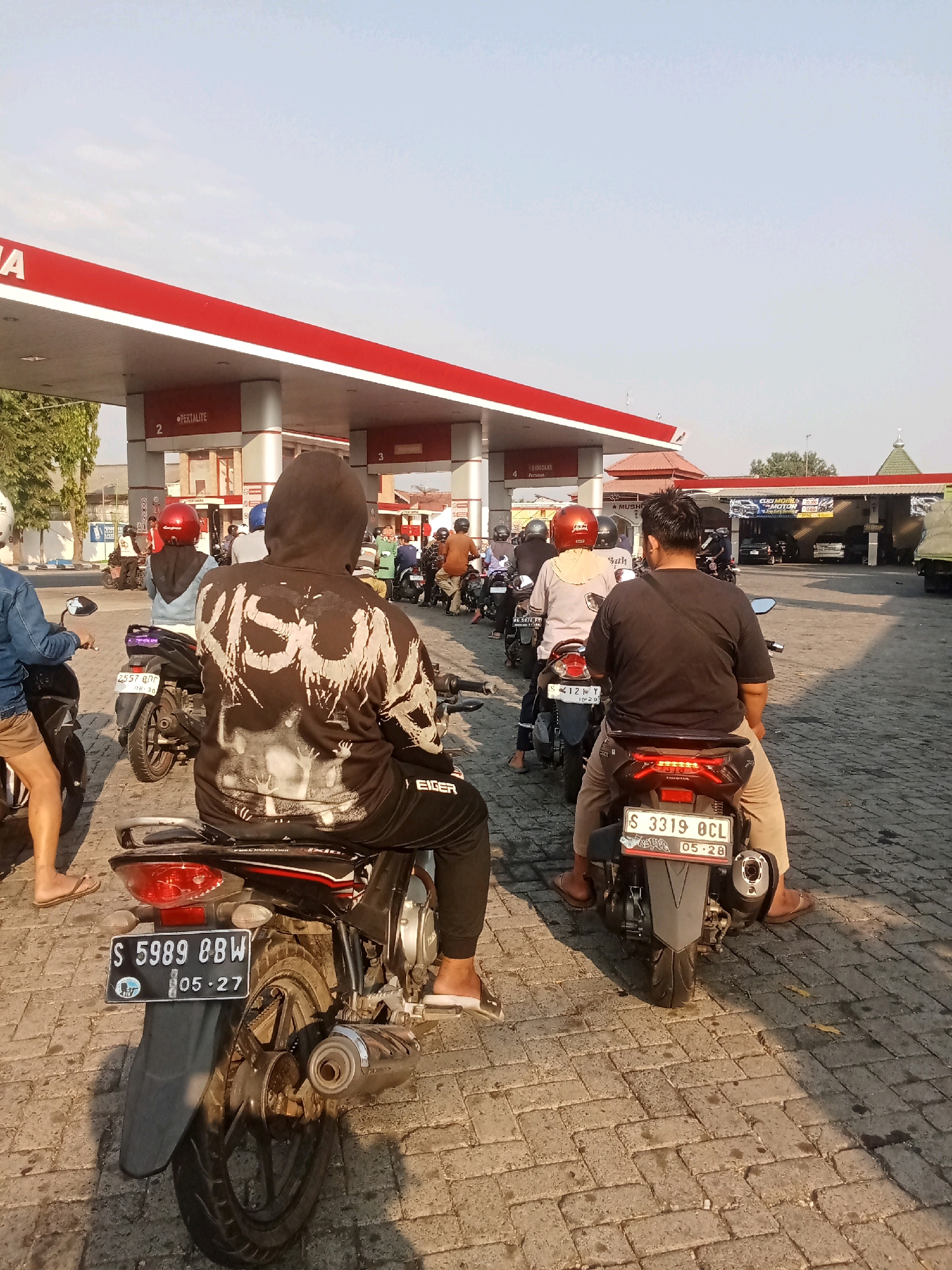 The image depicts a queue of motorcycles at a gas station in Jelakombo, Indonesia. The gas station features red and white signage, with the brand name "PERTAMINA" visible. Several individuals are seated on their motorcycles, facing away from the camera, waiting to refuel. The ground is paved with bricks, and sunlight casts shadows across the scene. In the background, other vehicles, including cars and a truck, are also present at the station. The sky is clear and blue, suggesting fair weather.