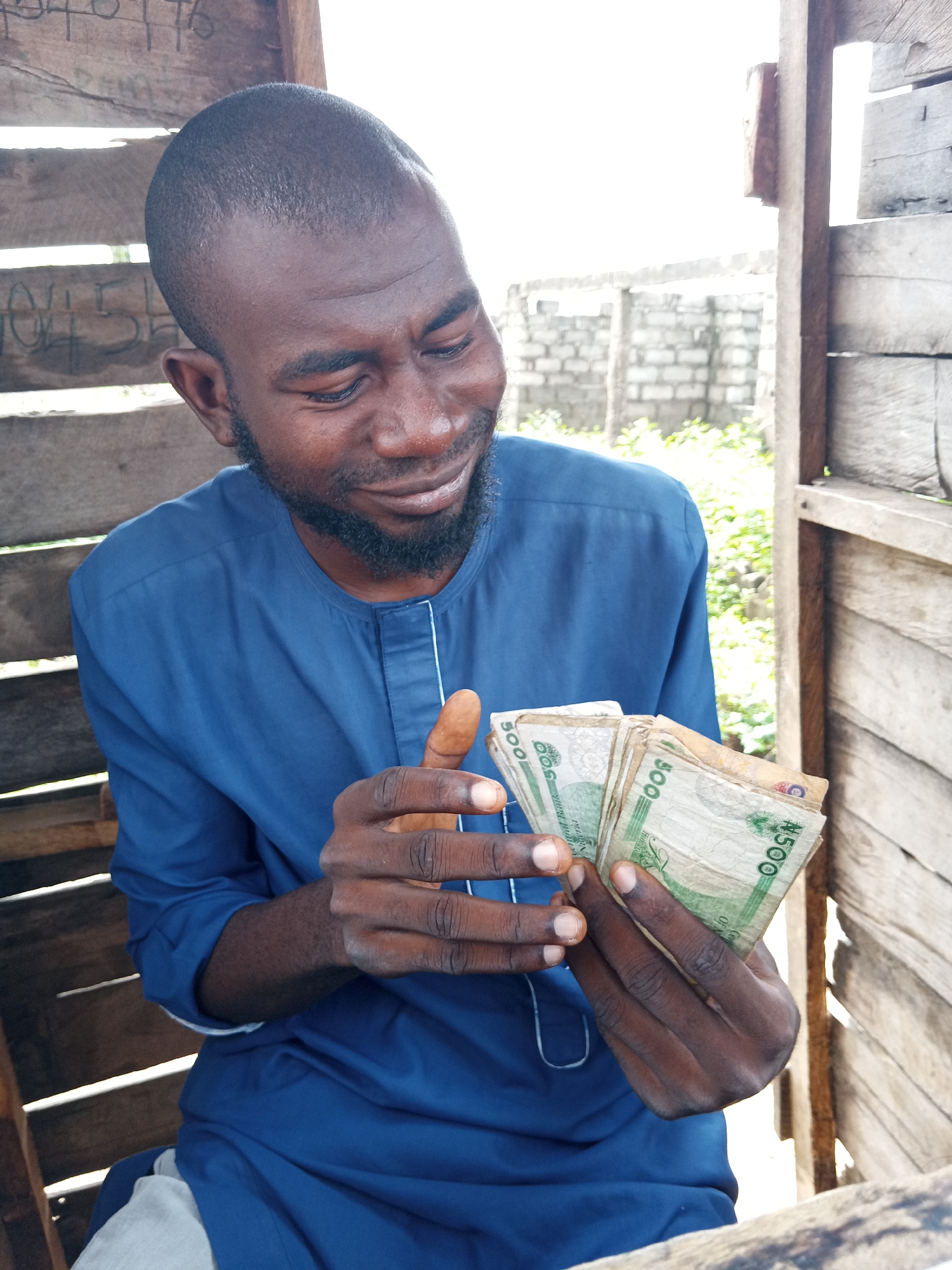 A man in a blue shirt is counting a stack of 500 Naira notes in a wooden structure. The location is the city of Unknown, Unknown. The man is looking down at the money with a slight smile. The wooden structure is made of planks and appears to be a small shack or stall. In the background, there is a partially constructed brick building and some greenery. The scene is captured in daylight with natural lighting.