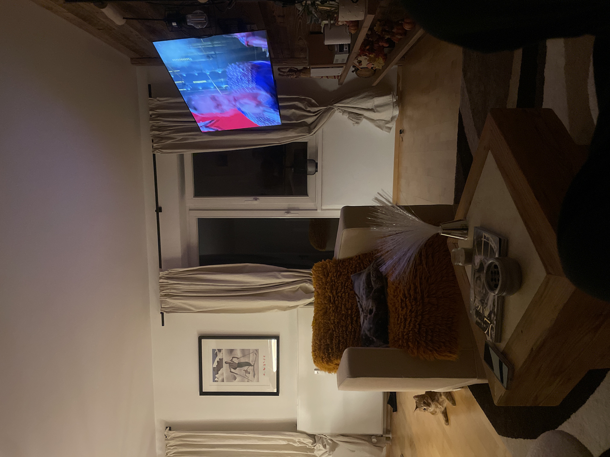 Here is a detailed description of the image:
**Setting:** The photo appears to be taken indoors, within a residential living room. The location is Unknown, Unknown, but no information about the specific city is known.
**Environment:** The room has off-white walls, with a large window positioned between the curtains. A TV is wall-mounted above the window.
**Objects:**
- Furniture: A light-colored armchair with orange, shaggy throw pillows is visible. There is also a wooden coffee table with a few items on it, including what appears to be magazines and decorative containers.
- Décor: A framed picture hangs on the wall below the window. Other décor is present on a shelf above the TV.
- Technology: A television screen displays content.
- Miscellaneous: Fiber optic lamp.
**Subjects:**
- An orange cat appears to be near the armchair.
- A person, only partially visible, is close to the coffee table.
**Actions/Interactions:** The interaction is implied, with the person likely watching TV.
**Essential Details:**
- Lighting: The room is illuminated by a combination of natural light coming through the window and artificial light sources.
- Composition: The photo is shot from a high angle.
