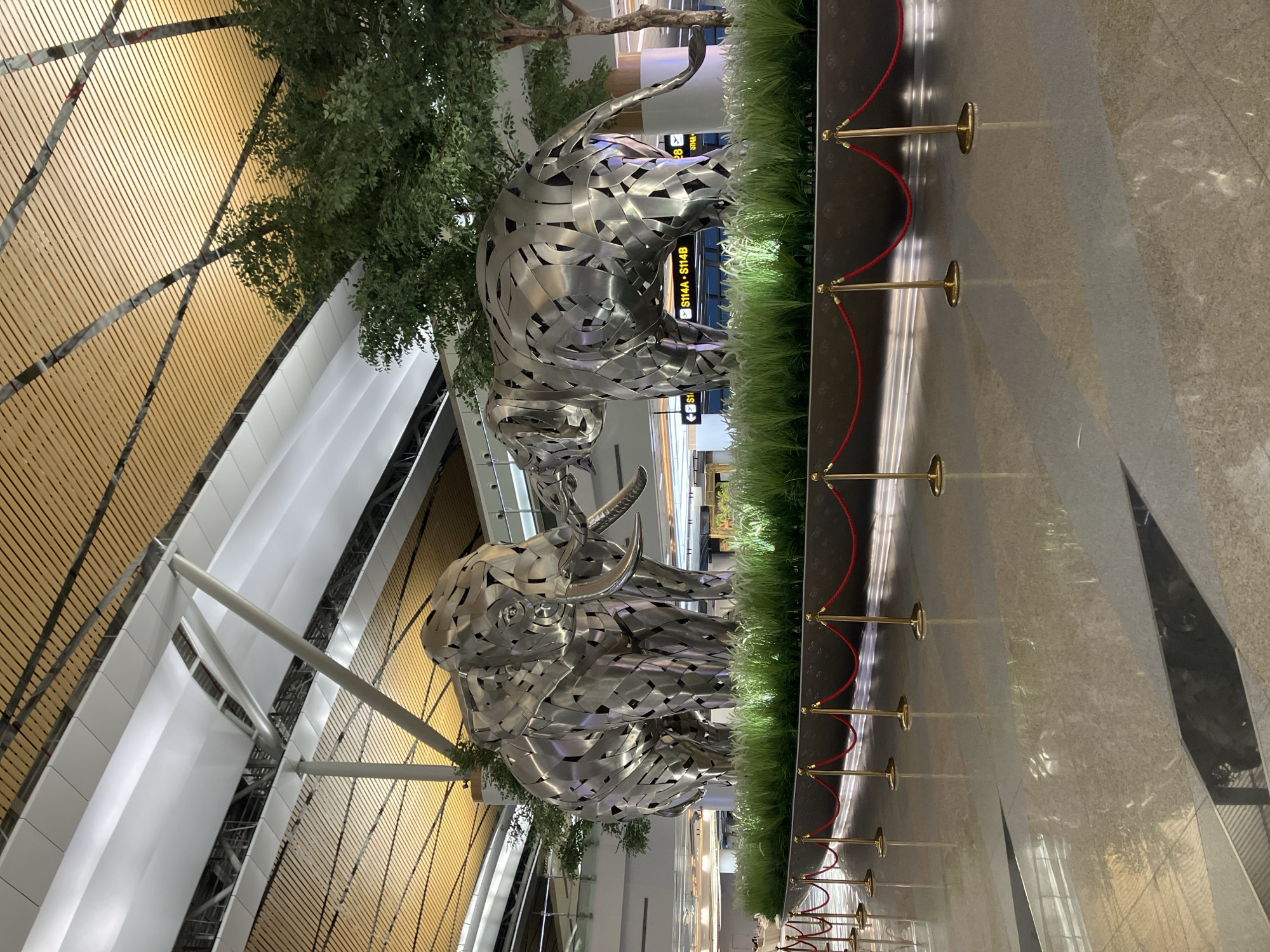 The image depicts two metal elephant sculptures in what appears to be an airport or large indoor public space. The sculptures are constructed from interconnected metal pieces. They are positioned next to a long planter with artificial grass, lined by gold-colored stanchions and red ropes. Behind the sculptures is a tall tree. The architecture includes a light-colored wooden ceiling and white paneling. Signage in the background suggests an airport terminal setting (with flight information). The reflective floor and overall aesthetic indicate a modern, well-maintained environment. The location is Nong Prue Subdistrict, Thailand, as it is a large public area in Thailand with the metal sculptures.