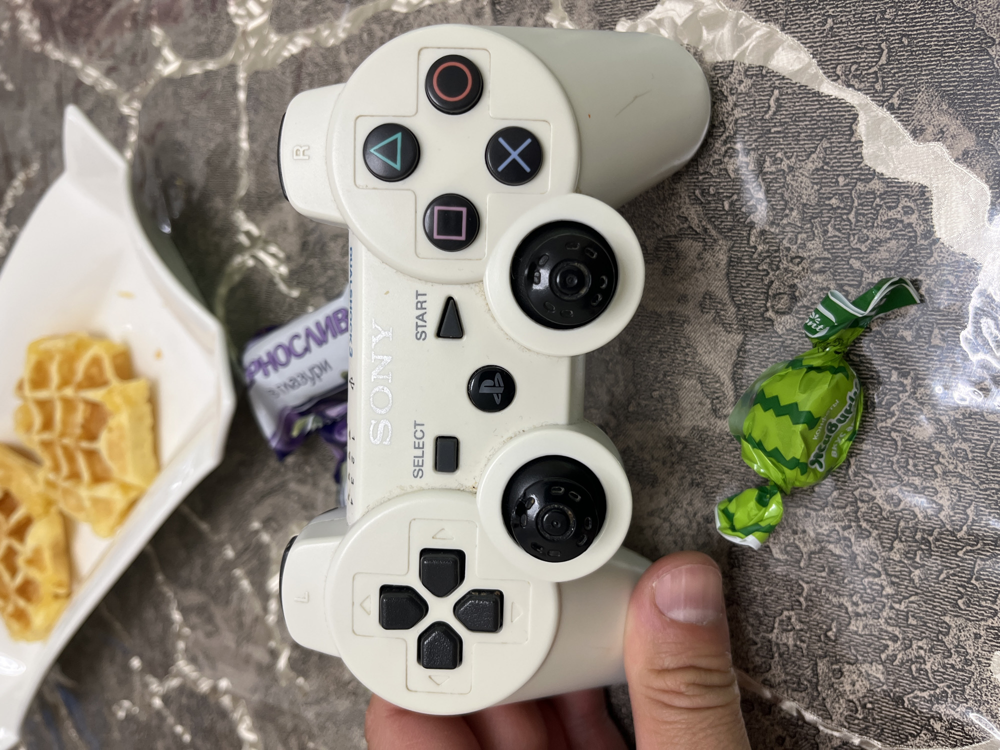 The image shows a white PlayStation 3 DualShock controller held in a person's hand, positioned on a textured surface. To the left of the controller is a white dish containing waffle pieces. A candy wrapper partially hides behind the controller. There is also a piece of candy in a watermelon-patterned green wrapper beside the controller. The city of Sochi, Russia is not identifiable within this image.