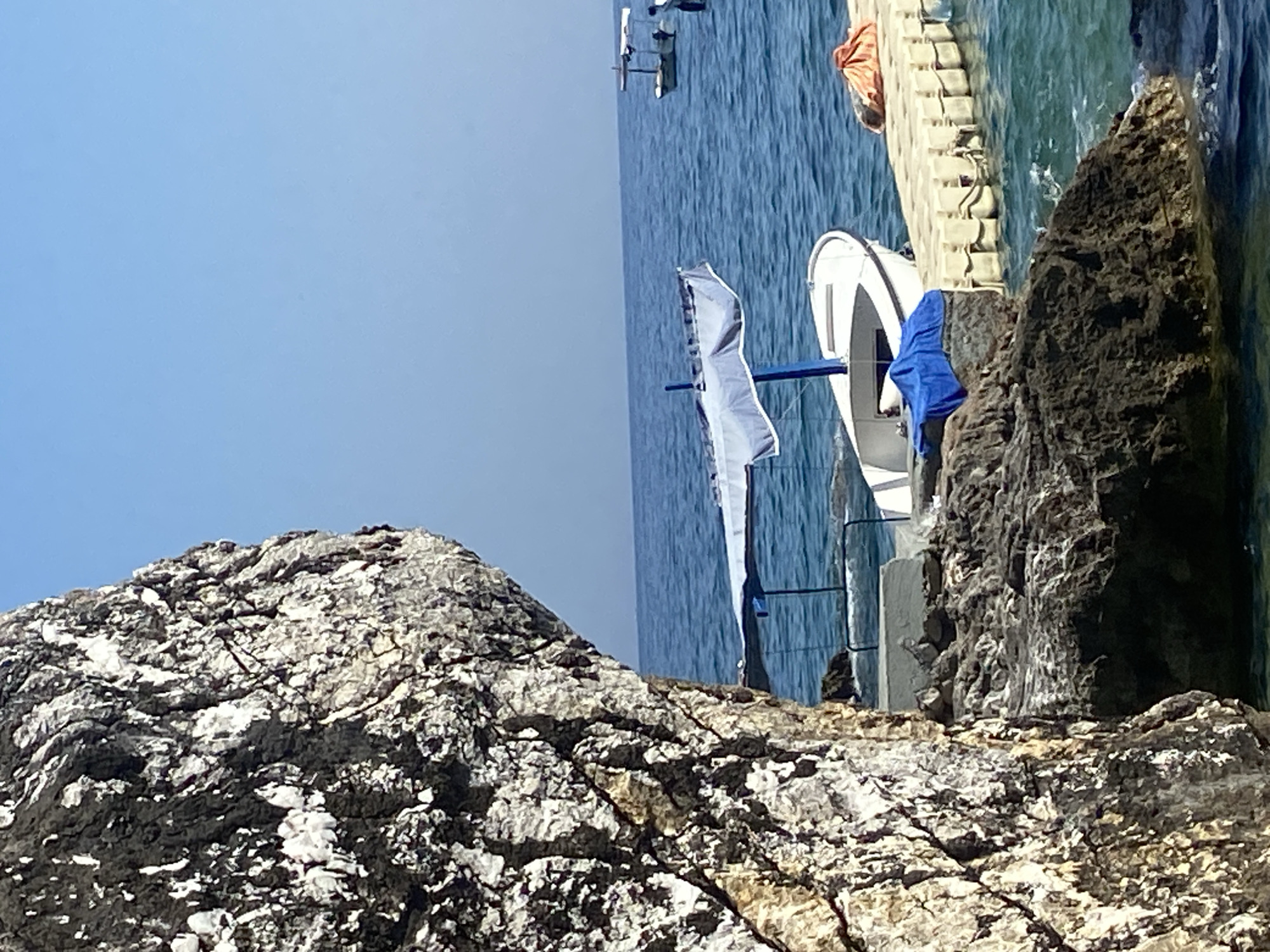 In Prijevor, Montenegro, a bright, sunny day unfolds by the sea. A large, textured rock formation dominates the left foreground. Beyond it, the azure water of the Adriatic Sea stretches to the horizon under a clear blue sky.
A white boat with a blue canopy is moored near the shore, with a blue cloth draped over its bow. Adjacent to the boat is a modular plastic pontoon dock, beige in color, with an orange bundle resting on its surface. In the distance, another small boat with a canopy is visible on the water. The scene suggests a tranquil coastal setting.