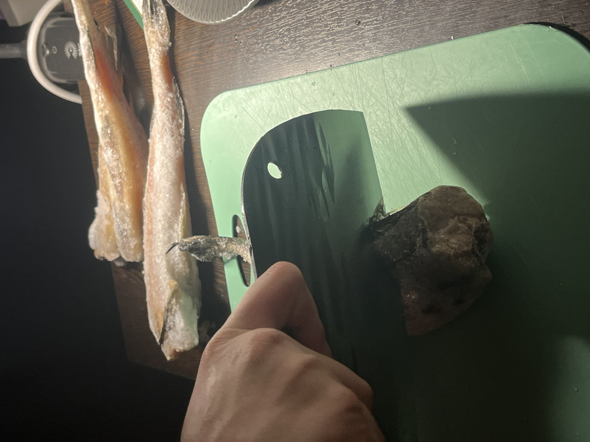 The image shows a close-up, oblique overhead shot of a food preparation scene. A light green cutting board sits on a dark, possibly wooden, surface. On the left, three frozen fish are positioned, while a large cleaver is cutting through a gray, possibly frozen, food item. A human hand is holding the cleaver. The scene appears to be set indoors, possibly in a kitchen, given the presence of the cutting board and food items. There is nothing to indicate location in the city of Saint Petersburg, Russia.