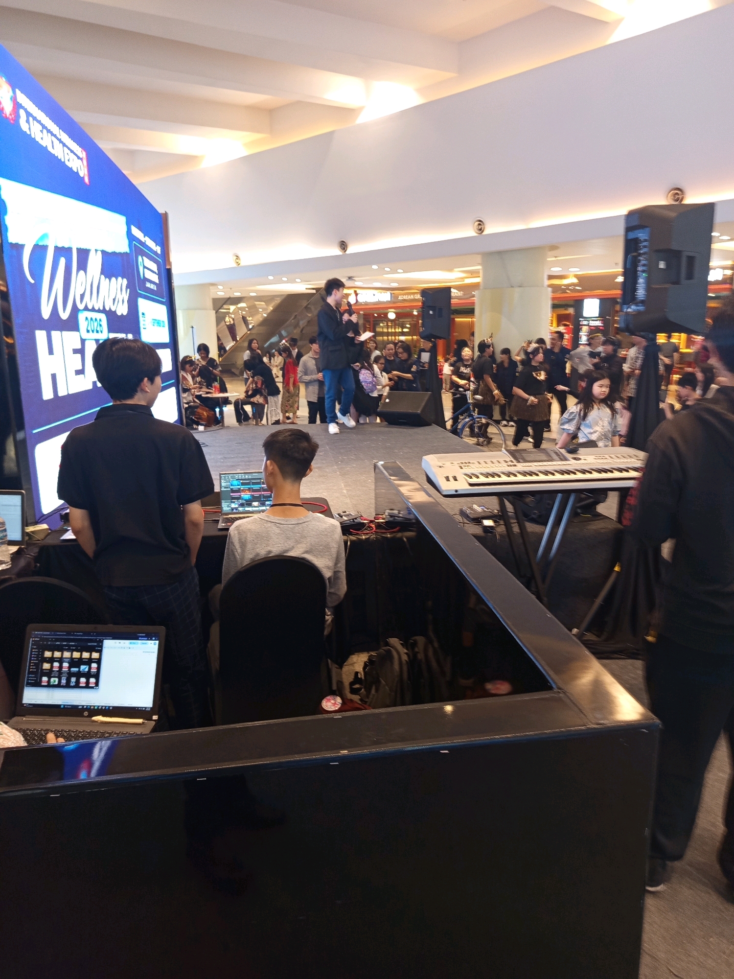 The image depicts an indoor event, likely a mall or exhibition center, in West Jakarta, Indonesia. A large blue digital screen displaying "Wellness 2025 HEALTH EXPO" is prominently visible on the left. In the foreground, two young men are positioned behind a black counter, one wearing a black polo shirt and plaid pants, the other a gray t-shirt. The man in gray is seated, facing a laptop with an audio editing software open. The man in the black shirt stands and looks towards the audience.
On a raised stage in the mid-ground, a young girl with long dark hair is seated at a white electronic keyboard. Behind her, two men in dark clothing stand near a microphone and a large speaker. The background is filled with a crowd of diverse individuals, suggesting an active event with attendees milling about and interacting. The overall environment appears to be a public space with elevated ceilings and various commercial establishments visible in the distance.