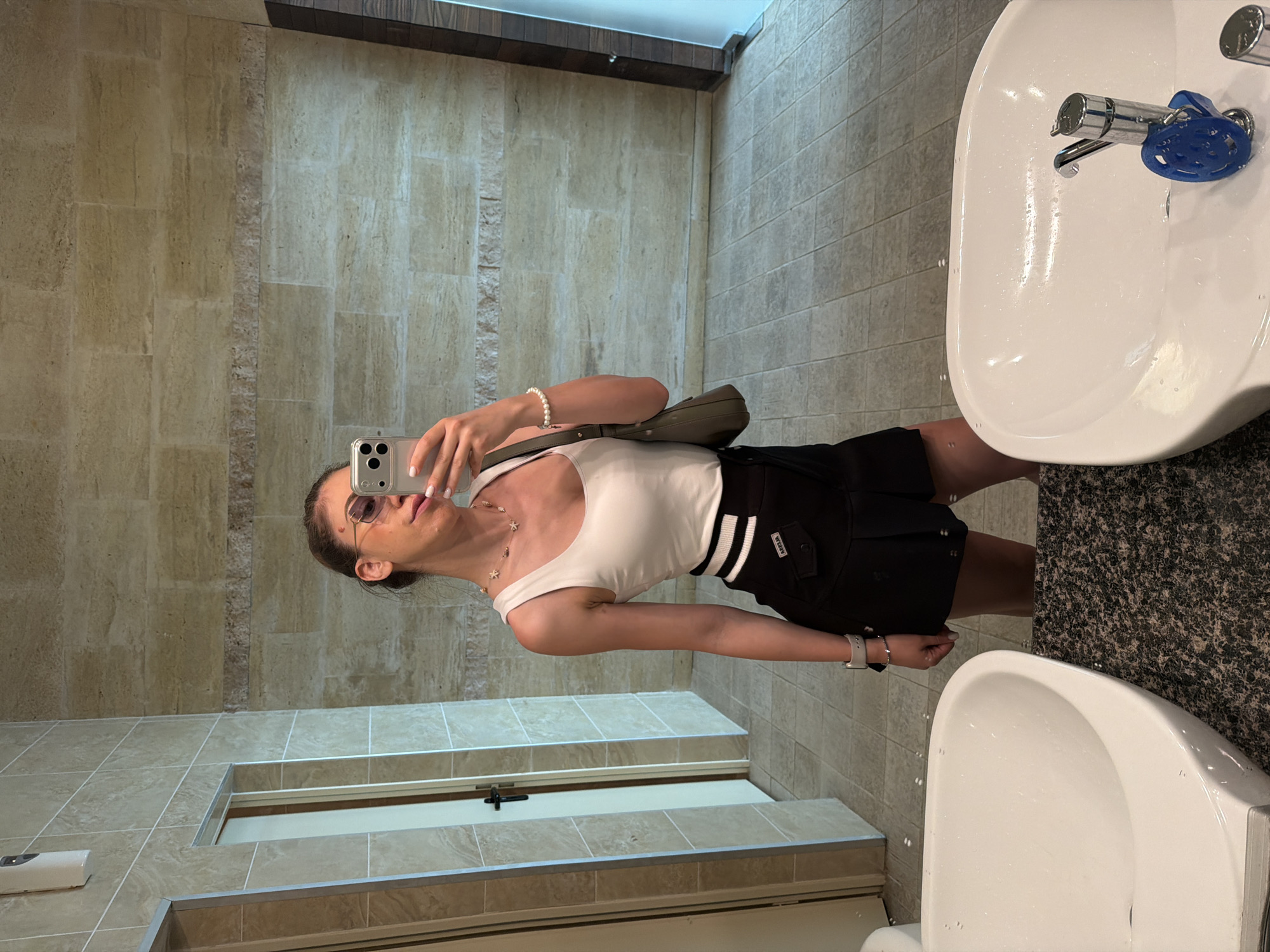 A young woman stands in a brightly lit restroom, taking a selfie in a large mirror. She is wearing a white tank top, a black skirt with white stripes, and sunglasses. She is holding a smartphone in her right hand and has a small green purse slung over her left shoulder. Her hair is pulled back in a bun, and she is wearing a pearl bracelet on her left wrist. The restroom has tiled walls and floors, and two white sinks are visible in the foreground. The scene suggests a public restroom, possibly in a hotel or a shopping center, given the clean and modern design. The image does not contain any specific visual cues to pinpoint the location to Primorsko, Bulgaria.