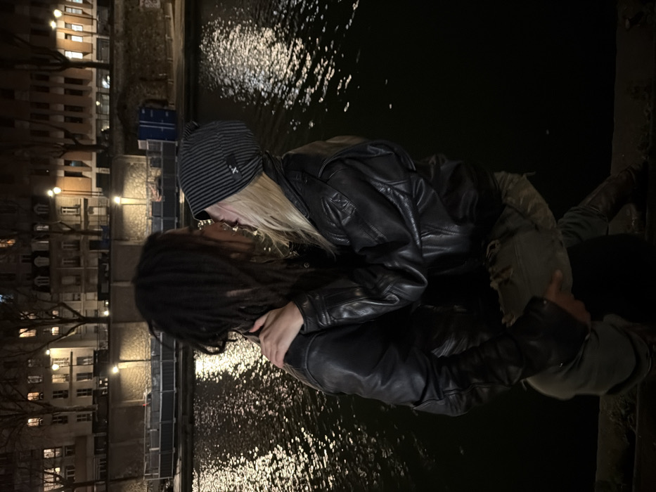 Two individuals embrace by a river at night. The person on the left has dark dreadlocks and a leather jacket. The individual on the right wears a striped beanie, a leather jacket, and has long, light-colored hair. They appear to be kissing. The river reflects light, and buildings with illuminated windows line the opposite bank.