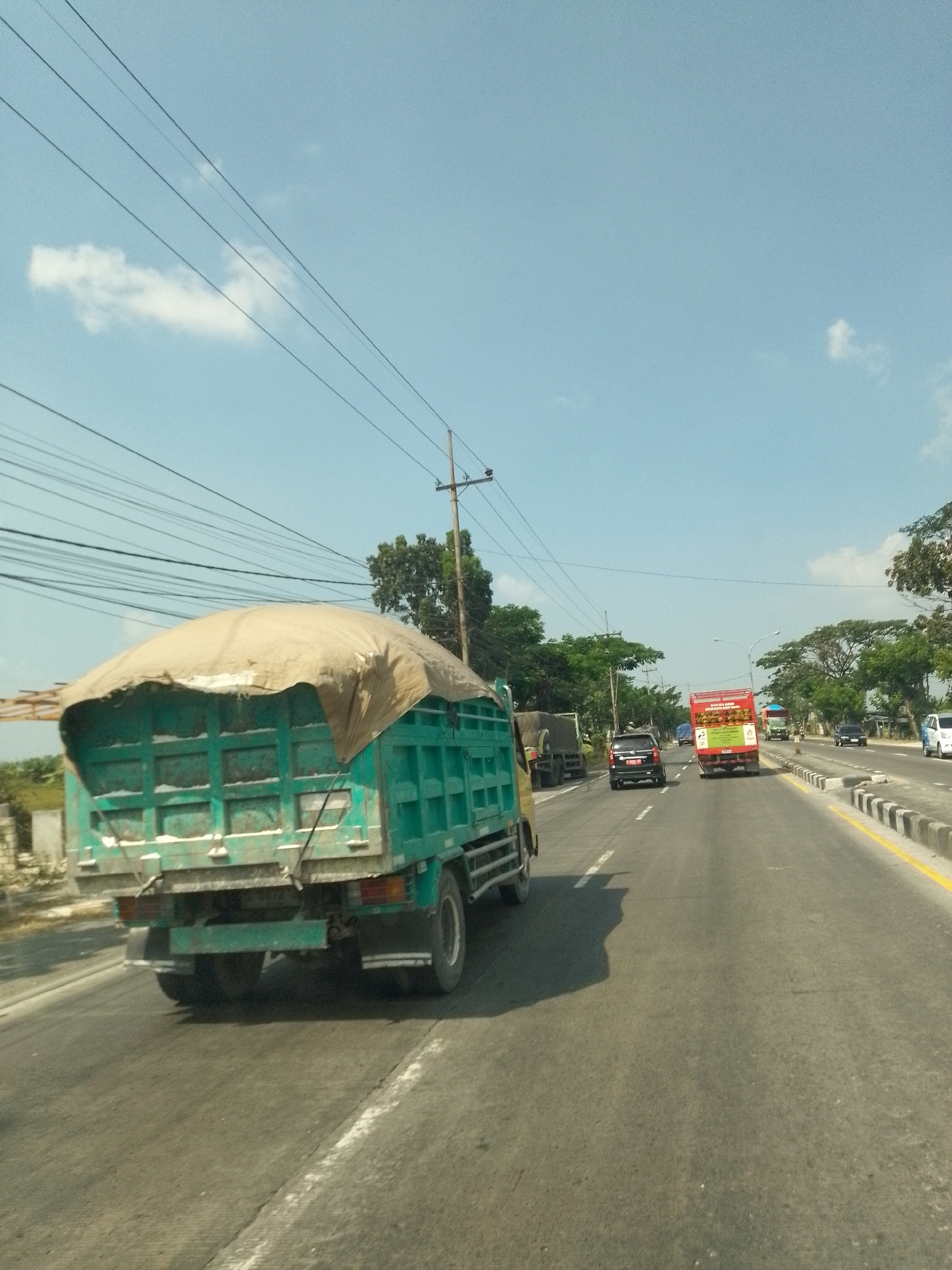 The image depicts a street scene in Waruwetan, Indonesia, under a bright, partly cloudy sky. The primary focus is a turquoise truck with a tarp-covered bed, seen from a rear-quarter angle, traveling on a paved road. The truck's tarp is a light brown or beige color, and it appears to be secured over the cargo.
Several other vehicles are visible on the road. Ahead of the turquoise truck, a red truck with advertising on its side is also moving. Further in the distance, there are other vehicles, including at least one car and what appears to be another truck. The road has multiple lanes marked with white and yellow lines.
The environment includes a clear sky with thin, elongated clouds. Power lines are strung across the upper portion of the image, supported by utility poles. Trees line the side of the road, creating a green backdrop in some areas. The road itself shows signs of wear, with some lighter patches and minor debris. The overall impression is of traffic on a highway or main road on a sunny day.