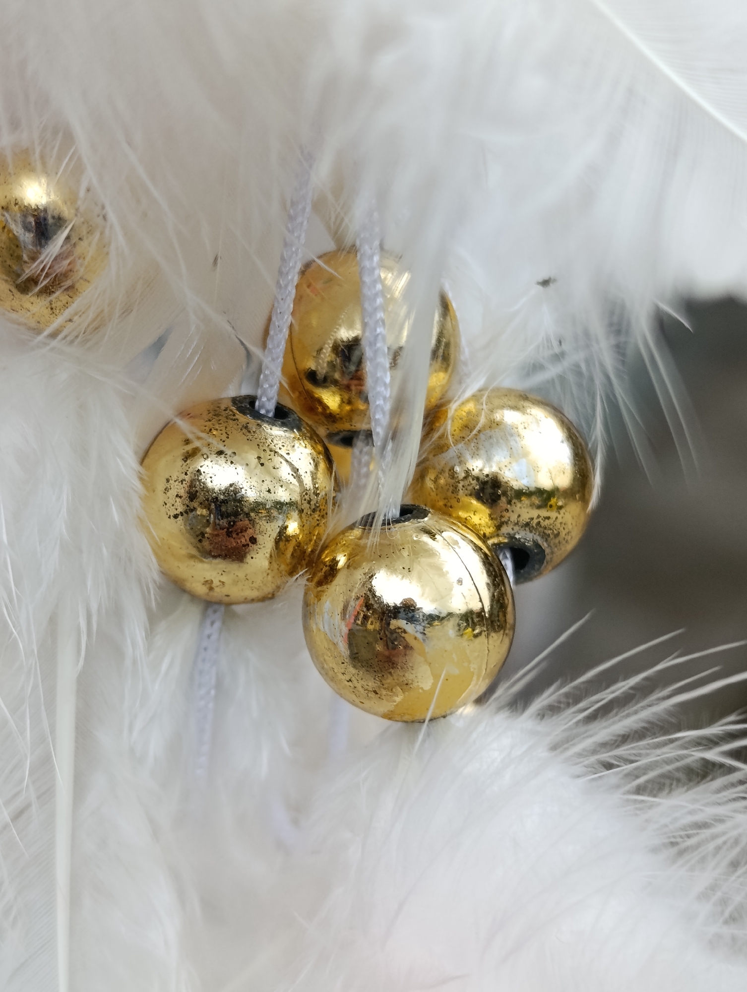 This is a close-up, indoor, high-angle, medium shot of a decorative arrangement. The subjects are several small, round, gold-colored beads, some with a speckled texture, strung on thin white cords. These beads are nestled within a fluffy, white material that appears to be feathers or faux fur. The background is softly blurred and appears to be a neutral gray. The lighting is bright and reflective, highlighting the metallic sheen of the beads. The image does not provide any indication of the location being Manali, India, or any specific urban setting. There are no people, actions, or interactions depicted.