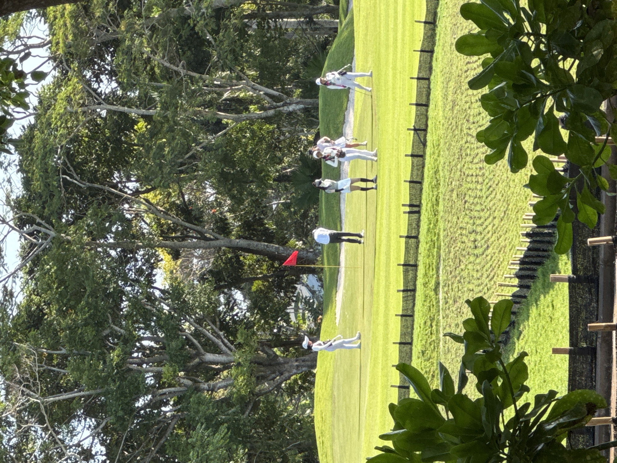 The image captures a scene on a golf course, likely in Kuta Selatan, Indonesia, given the tropical foliage in the foreground and background. A group of at least six individuals, presumed to be golfers, are dispersed across a manicured green. The people are primarily wearing white attire. A red flag marks the location of a hole. A tree line is prominent on the left, while a fence is on the right of the frame.