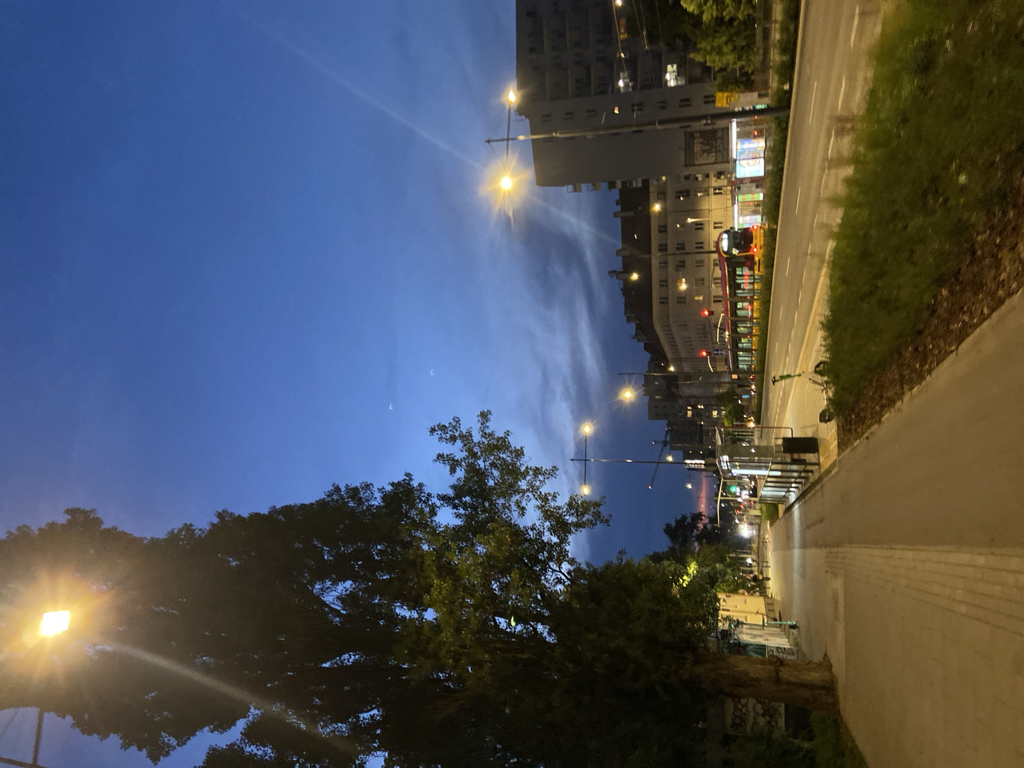 This is a nighttime street scene in Warsaw, Poland, likely in the Praga district, judging by the architecture and the presence of a tram. A wide, paved sidewalk runs along the left side of the image, bordered by a large, leafy tree. Streetlights cast a warm glow, illuminating the sidewalk and casting long shadows. Beyond the sidewalk is a multi-lane road with a tram visible in the distance. The tram is red and yellow and appears to be in motion. Buildings line the opposite side of the road, with some windows lit. The sky is a deep blue, transitioning to a lighter hue near the horizon, suggesting dusk or early morning. There are no people clearly visible in the image. The scene conveys a sense of urban evening activity.