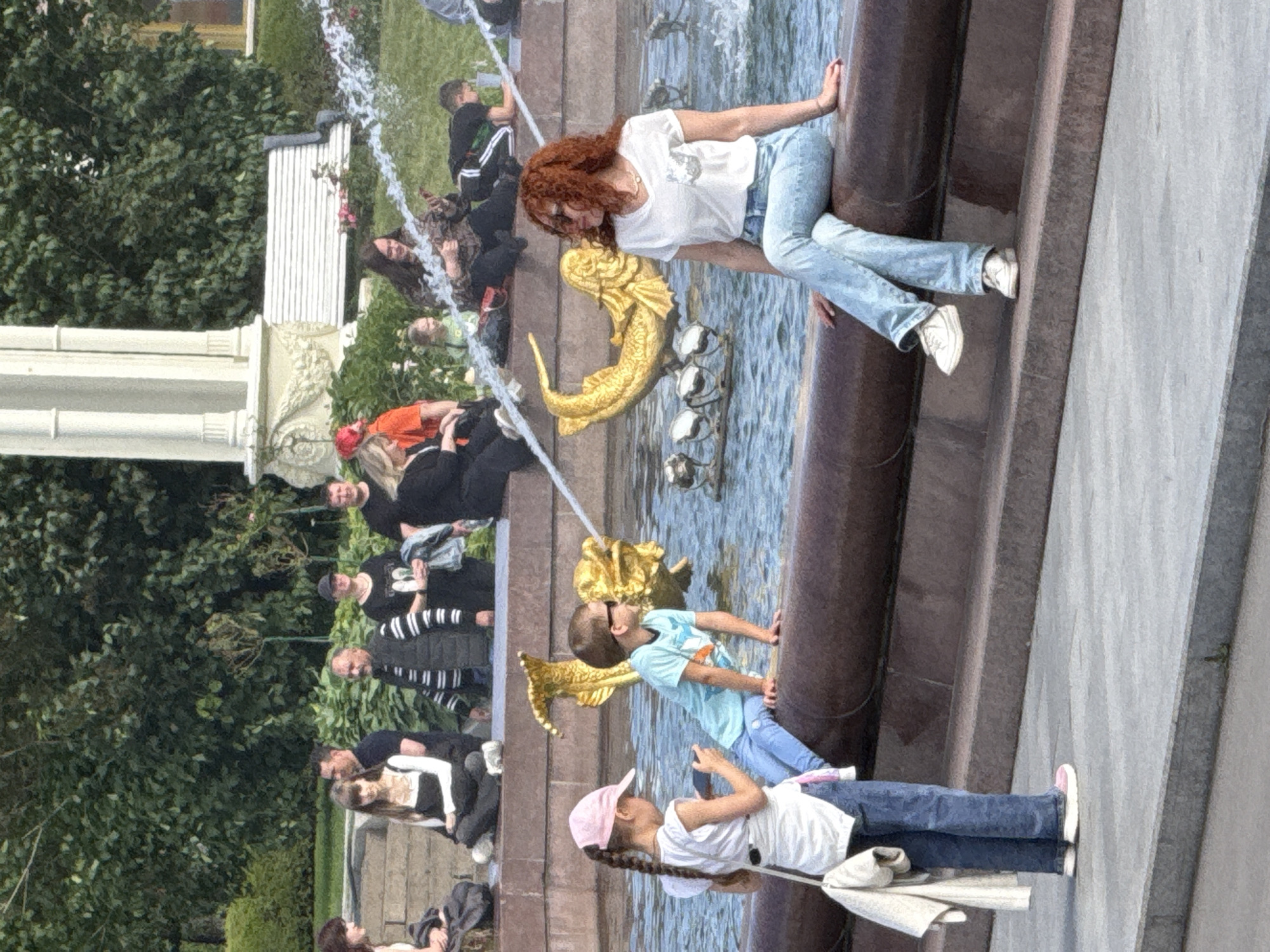 The image depicts a sunny day scene in a public park, likely the Alexander Garden in Moscow, Russia, given the architectural style of the white classical structure and the presence of a fountain with golden marine sculptures. In the foreground, a young girl wearing a pink baseball cap, white t-shirt, and blue jeans stands, holding a smartphone and appearing to take a picture. Beside her, a young boy in a light blue t-shirt and blue jeans sits on the edge of the fountain, wearing sunglasses. To the right, a woman with bright red curly hair, wearing a white t-shirt and light blue jeans, is seated on the fountain's edge, looking to her left. In the background, several other individuals are visible, some seated on tiered stone steps, and others standing near a white bench and lush green trees. The fountain features water jets and decorative golden fish sculptures.