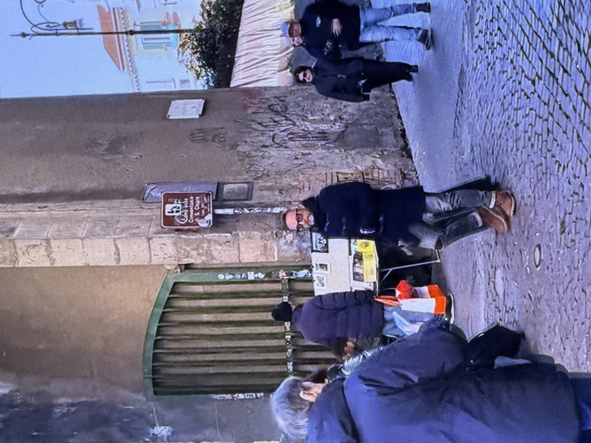 The image depicts an outdoor urban scene. Several individuals are visible, some walking and others standing. In the foreground, a person wearing a dark blue jacket is seen from the back, with their head turned to the left. To their right, another person in a dark coat and jeans is walking away from the camera. Further back, a person in a black hat and dark jacket walks past a green gate. A small table with various items, including what appear to be framed pictures, is set up near the gate. Several other people are in the background, some walking and some conversing. The environment includes a cobblestone street and a textured wall with some graffiti. A sign with text and a symbol is mounted on the wall. The overall lighting suggests it is daytime. There are no discernible elements in the image that place the scene in Dubai, United Arab Emirates.