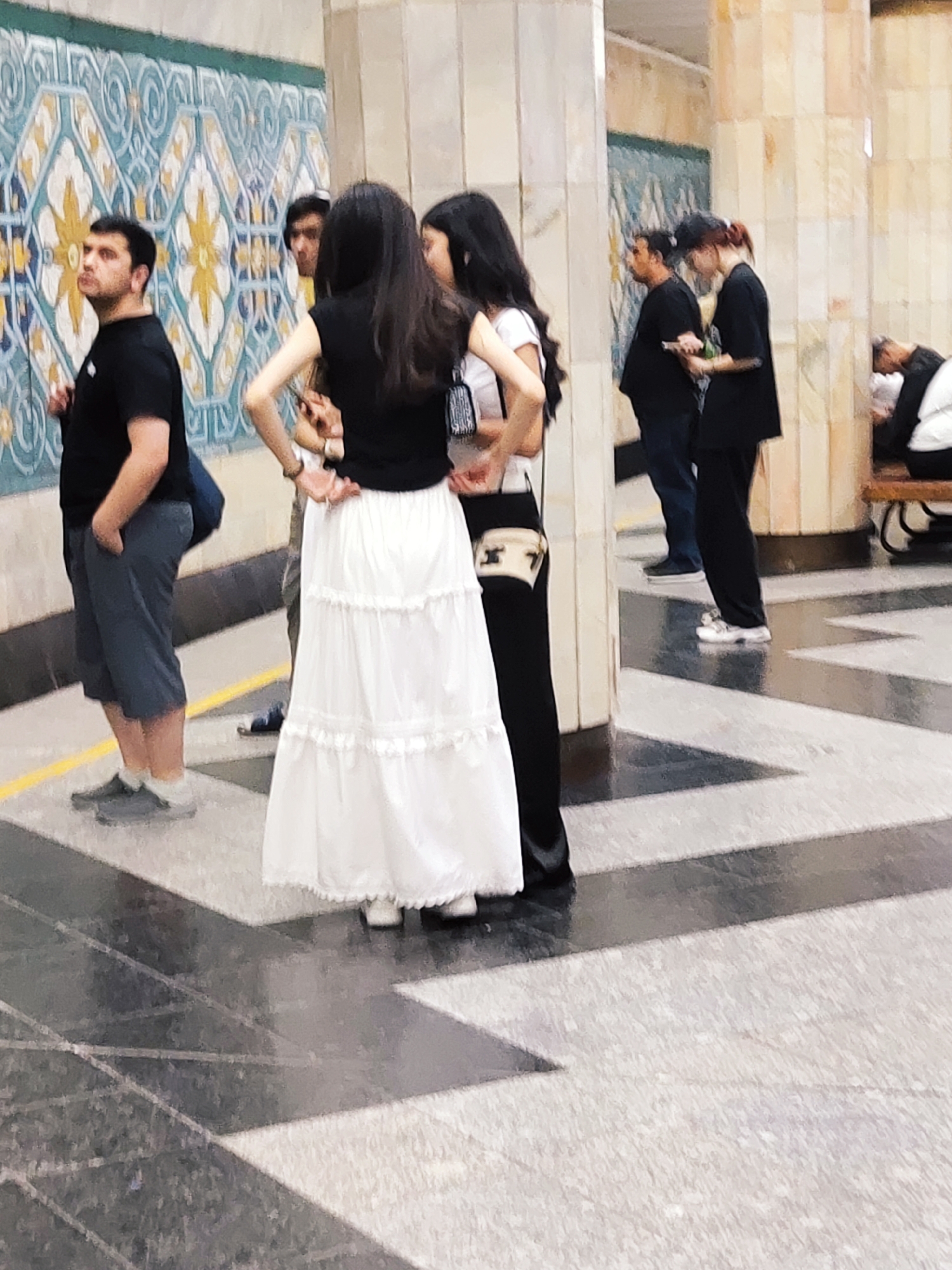 The image captures a group of people in a brightly lit, indoor public space with distinctive tilework on the walls and floor. The wall features a repeating pattern of blue, yellow, and white geometric shapes. The floor is a combination of dark and light gray patterned tiles.
On the left, a man in a black t-shirt and grey capri pants stands with his hands in his pockets, facing left and looking upwards. Next to him, a person with dark hair, possibly a woman, wears a black top and a long, tiered white skirt, and is looking downwards. To their right, another person with dark hair, wearing a black top and white capri pants, is partially visible. Further to the right, two individuals are interacting; one is wearing a black top and dark pants, while the other, with red-tinted hair, is wearing a black t-shirt and dark pants. In the background on the far right, a person is seated on a bench, bent over.
The overall scene appears to be a metro station or a similar public transit area in Tashkent, Uzbekistan, given the distinctive architectural and decorative elements. The lighting suggests an indoor environment. The people are casually dressed and seem to be waiting or observing.