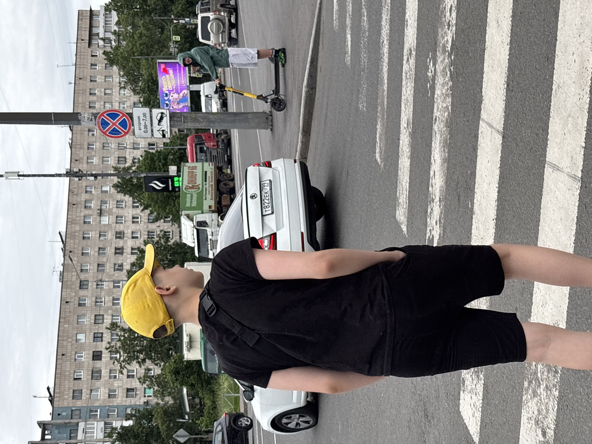 This image captures a street scene in Saint Petersburg, Russia, on an overcast day. In the foreground, a young person wearing a black t-shirt, black shorts, and a yellow baseball cap is standing on a pedestrian crossing. They have their hands in their pockets and are facing away from the camera, looking towards the street. A black strap is visible across their back, likely from a bag.
In the middle ground, a white Skoda automobile is parked on the left side of the road, facing away from the camera. To the right, a person wearing a green hooded jacket and white shorts is standing on a yellow electric scooter. This individual is also looking away from the camera, towards the street. Behind them, a traffic light pole with several signs is visible, including a red circular "no stopping" sign and a rectangular sign with Russian text and time information.
The background features a large, multi-story residential building with a grey facade and numerous windows. Trees and other vehicles, including trucks and SUVs, are also present, indicating an urban environment. The road surface shows clear markings for the pedestrian crossing. The overall atmosphere is neutral, reflecting a typical day in a city.