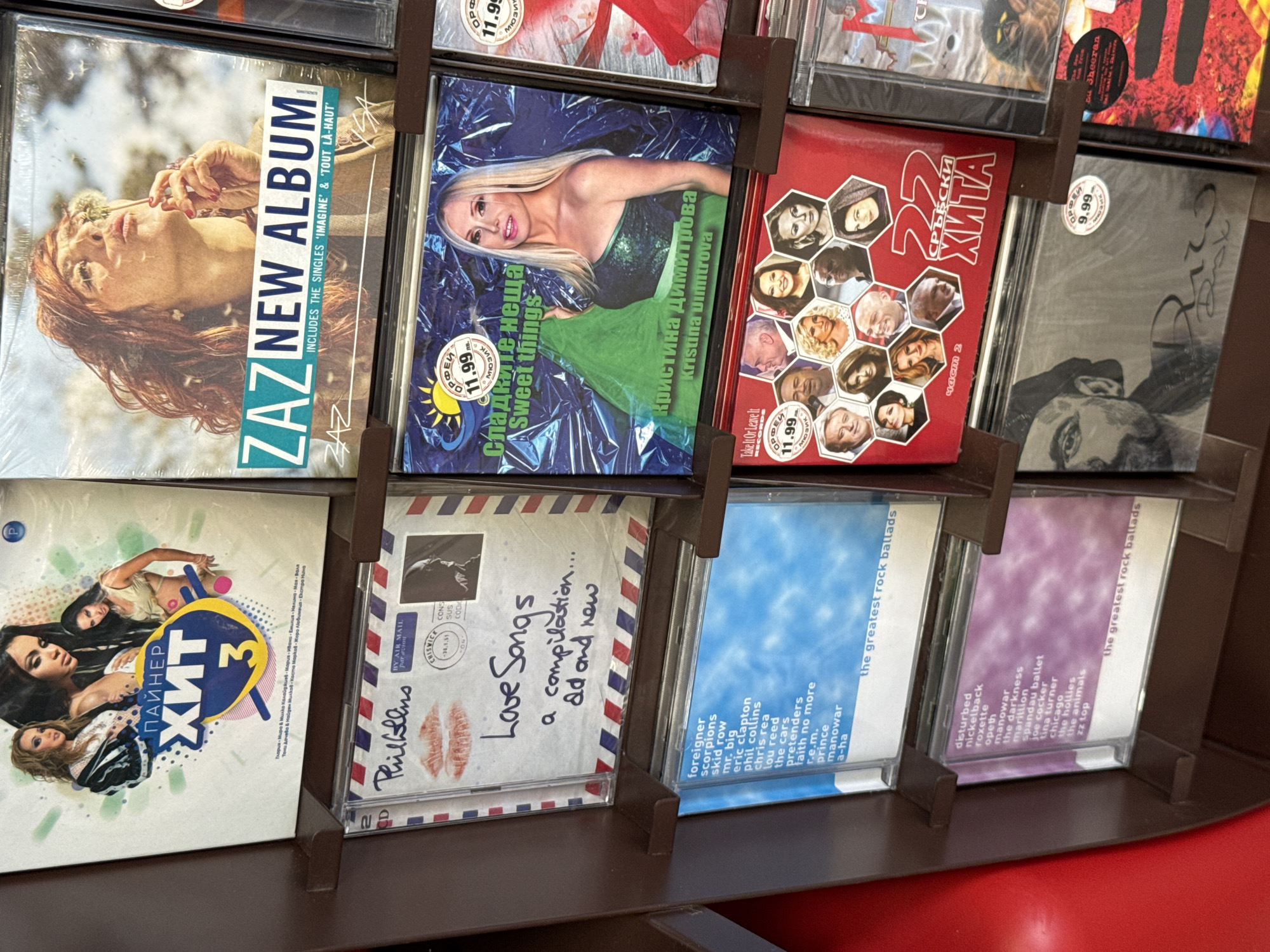 The image displays a retail shelf in Varna, Bulgaria, stocked with CDs. The focus is on the various album covers. Prominently featured are several compilation albums, including "Пайнер Хит 3," a ZAZ album titled "NEW ALBUM," and a CD labeled "Sweet Things" by Kristina Dimitrova. Other visible albums include "Love Songs - a compilation... old and new," and various "greatest rock ballads" compilations featuring artists like Foreigner, Skid Row, Eric Clapton, Phil Collins, R.E.M., Disturbed, Roxette, and ZZ Top. There is also a red CD featuring multiple portraits of people with the text "22 СРПСКИ ХИТА" and another CD with a sketched portrait and the name "Rico" visible. The lighting suggests an indoor environment, likely a music store.