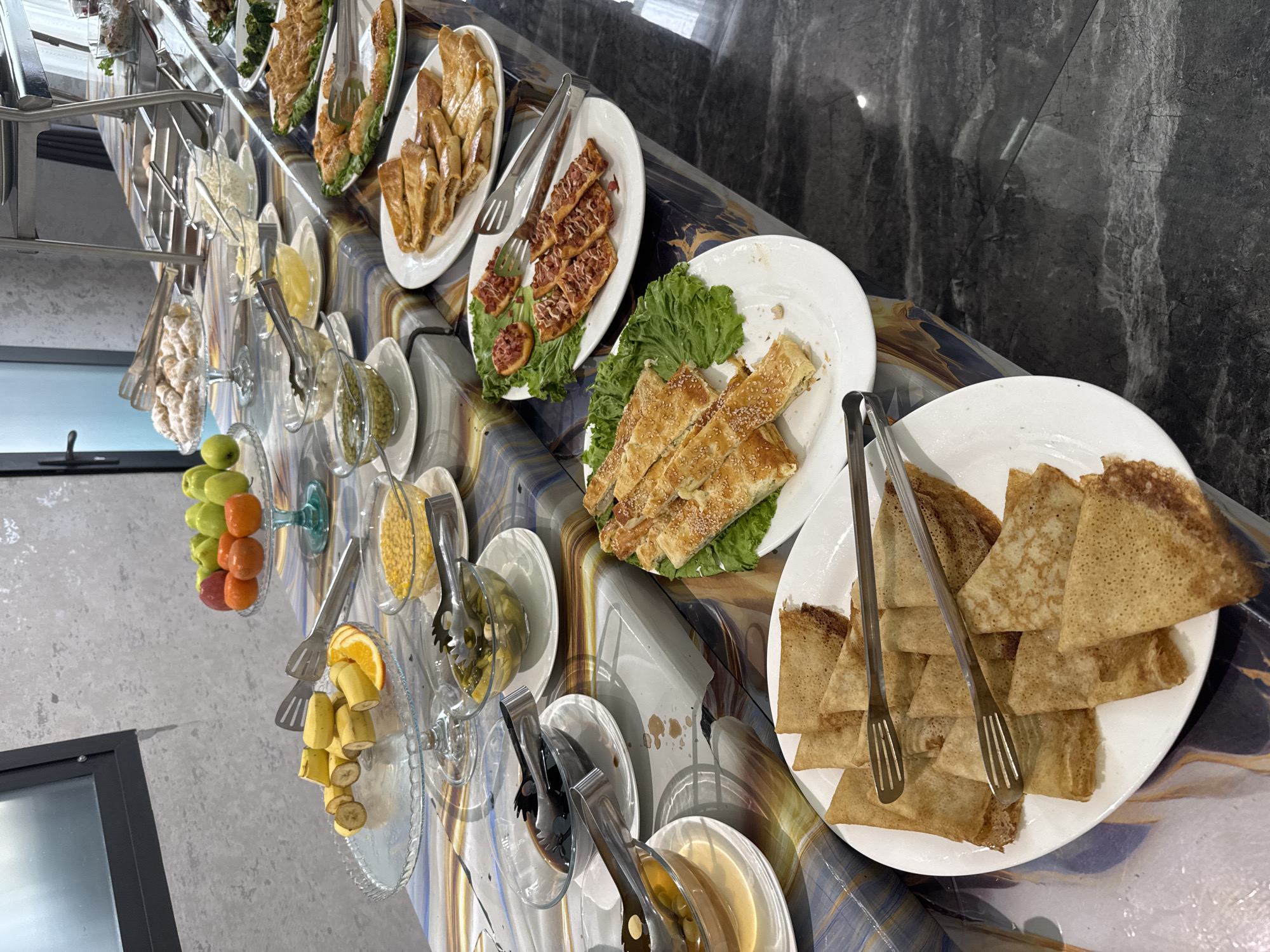 The image shows a buffet table in what appears to be an indoor dining area. The buffet includes a variety of food items, such as pastries, pancakes, and fruits, served on white plates and clear glass stands. Serving tongs are present next to each dish. The background is a marble-like wall. The context of the location being in Tashkent, Uzbekistan is not visually ascertainable from the image itself.