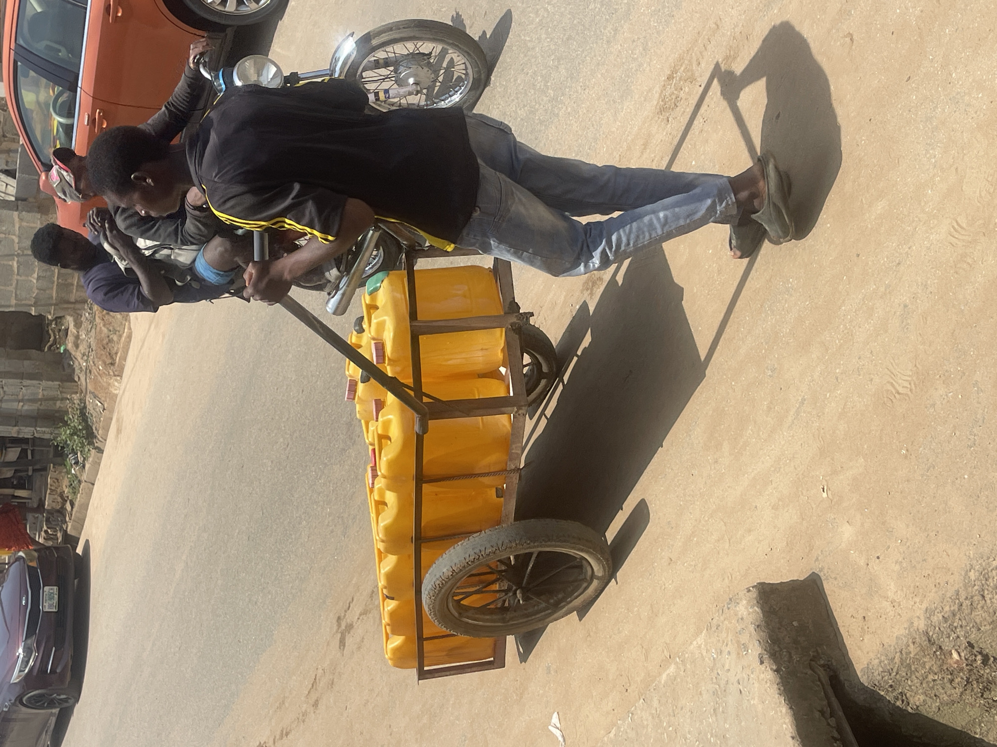 The image depicts an outdoor scene on a dirt road, likely in Abuja, Nigeria, judging by the context. The sun is high, casting strong shadows, suggesting it is midday. Two young men are visible. One, in the foreground, is pushing a cart loaded with numerous bright yellow plastic jerrycans. He wears a black t-shirt with yellow stripes on the sleeves and blue jeans. The other man, seated and partially obscured, is wearing a dark purple shirt and light blue shorts. The cart appears to be metal-framed with two wheels, one of which is prominently featured in the lower right quadrant of the image. Behind the men, an orange car is parked, and a motorcycle is partially visible. The background shows a dusty, uneven road surface and some structures that hint at a semi-urban or rural environment. The overall impression is of a hot, sunny day with people engaged in a task that involves transporting goods, possibly water or fuel, given the jerrycans. There is no visible text in the image.