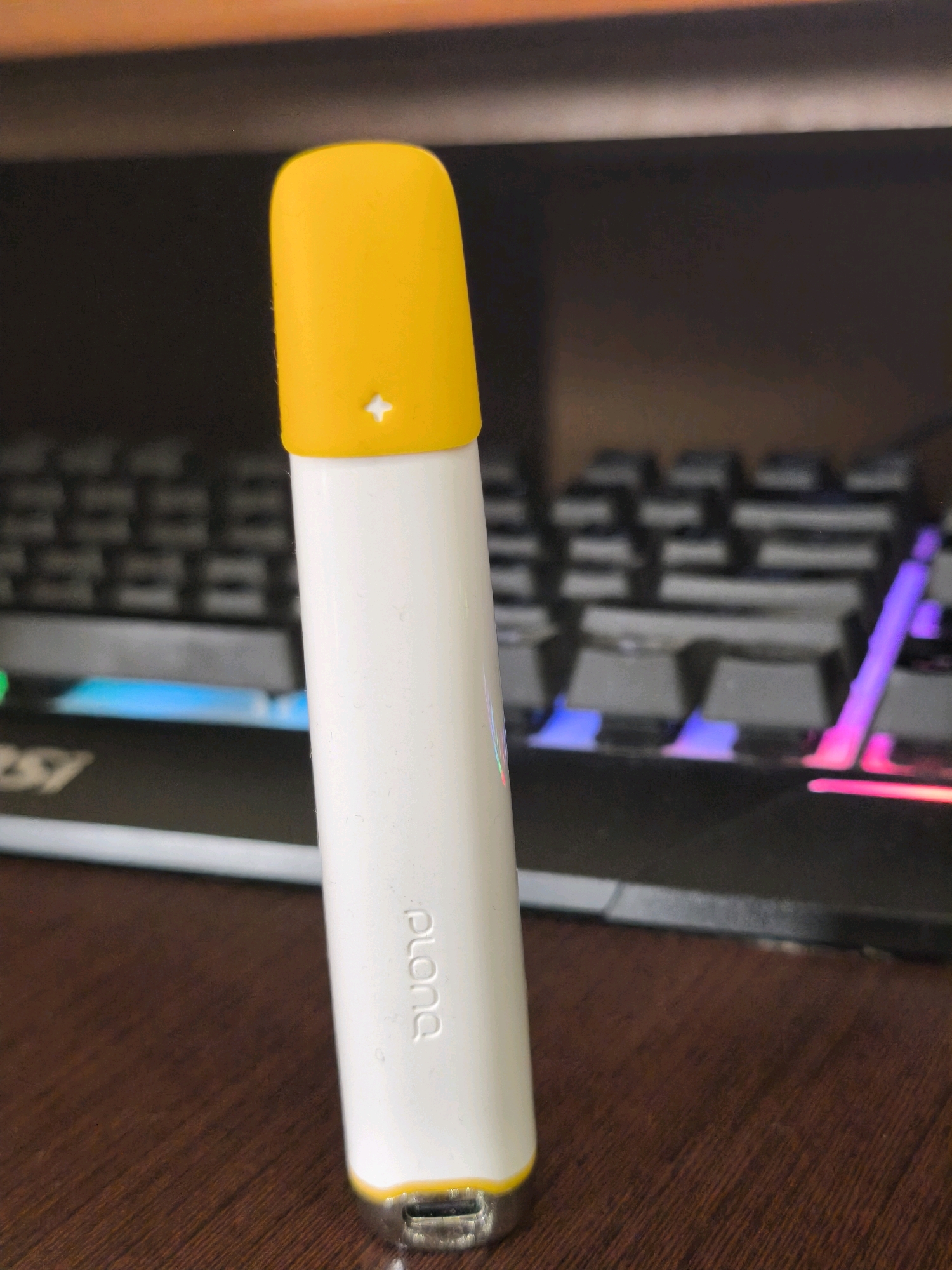 A close-up, eye-level, indoor shot features a white cylindrical device with a yellow cap and a metallic base, resting on a wooden surface. The device has embossed text that reads "plona" vertically. Behind the device, a keyboard with illuminated keys in blue and pink is visible, suggesting a desktop setup. The background is dark and out of focus. The scene appears to be a personal workspace. There is no indication of the city of Karachayevsk, Russia, or any people present.