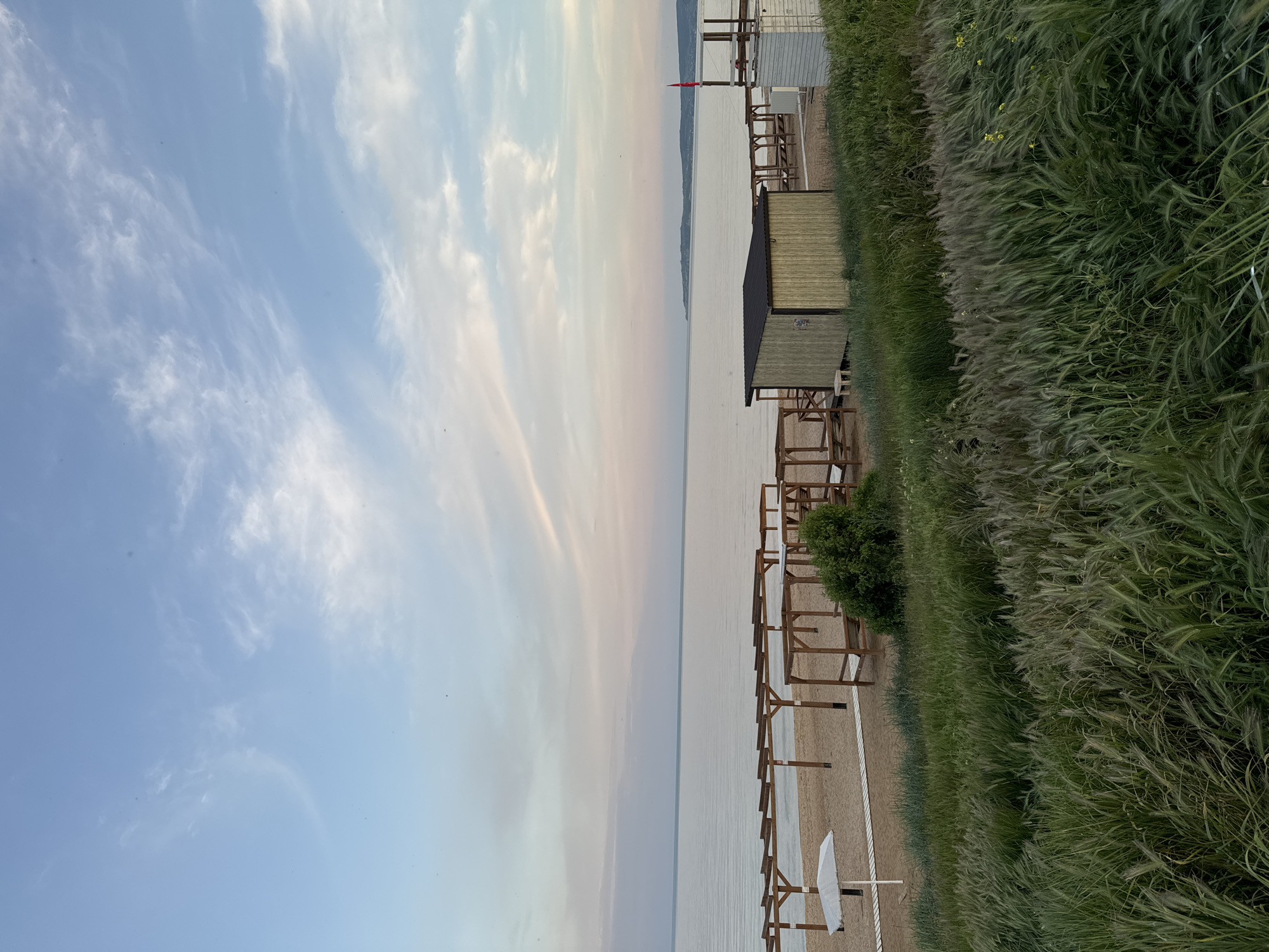 The image shows a beach in Береговой сельский совет, Ukraine. The view is from the shoreline, looking out over the water towards a distant landmass. The sky is light blue with scattered white clouds. On the right, there is a grassy area with tall, green vegetation. Wooden structures, possibly cabanas or shelters, line the beach, with some being covered and others open. The beach itself is sandy. The water is calm and reflects the sky's colors. A red flag is visible in the distance.