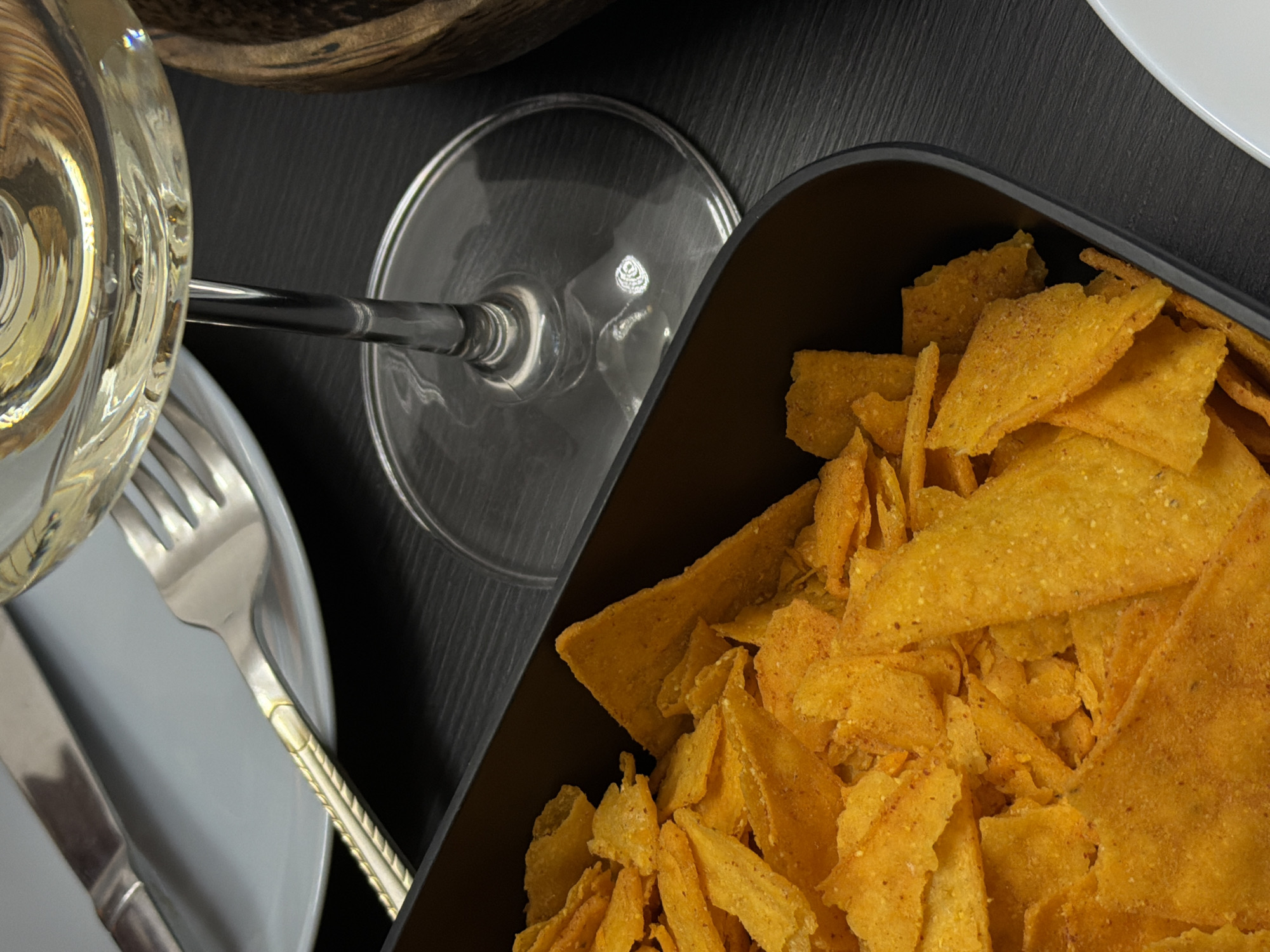 The image shows a close-up of a table setting. A clear wine glass filled with white wine is visible next to a plate with silverware (knife and fork). A black bowl filled with tortilla chips sits on the right side of the frame. The table surface is dark, possibly black slate. The overall impression is a meal setting, perhaps an appetizer course. Location information is not available in the provided image.